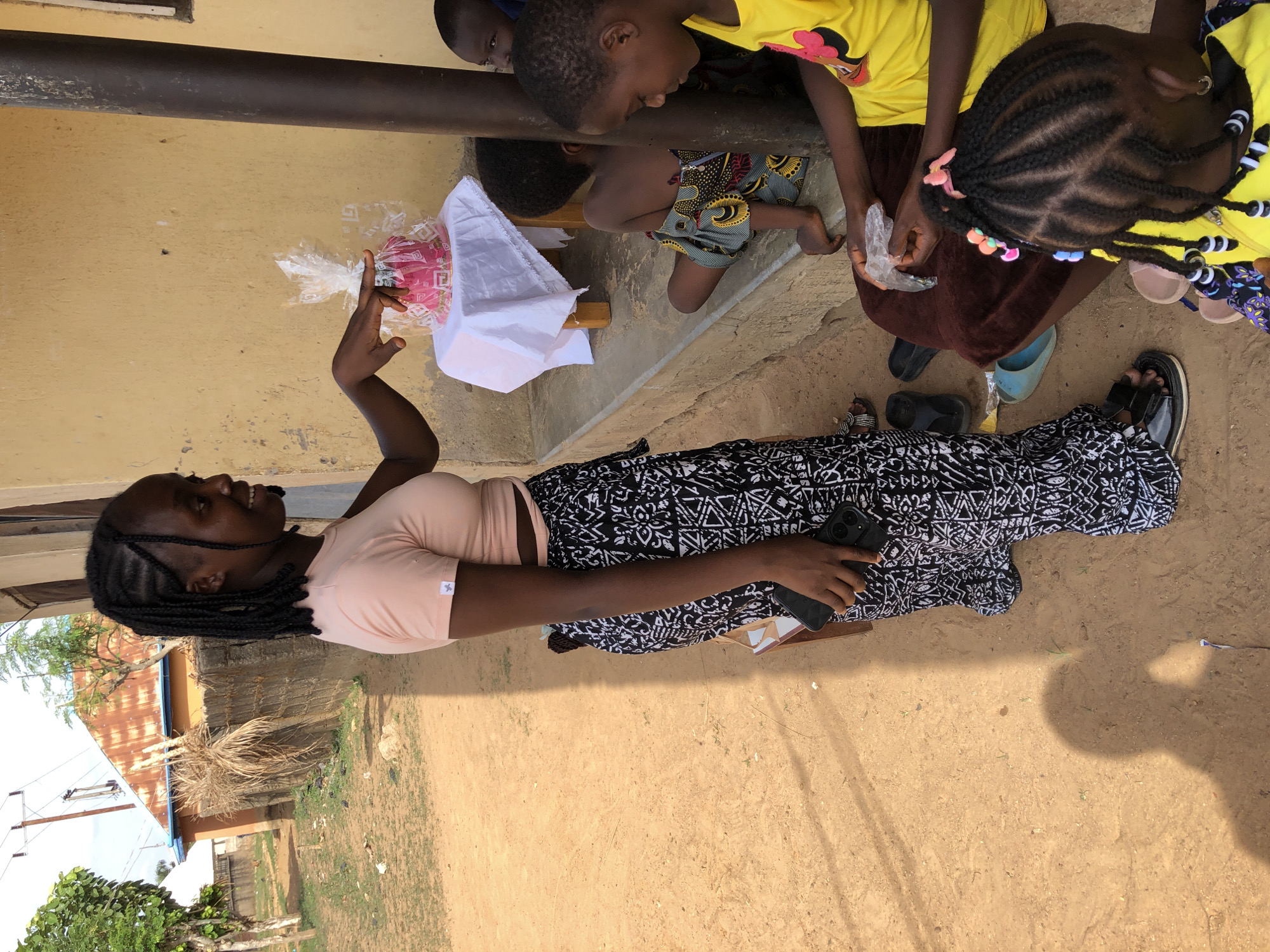 In Akwanga, Nigeria, a woman with dark skin and braided hair wears a peach-colored top and a black and white patterned skirt while sitting on the ground outdoors. She holds a black smartphone in one hand and gestures with the other towards a bouquet of pink flowers wrapped in clear plastic and white paper, which rests on a small wooden stool.
Three children stand near the woman. One child wears a yellow t-shirt, and another is dressed in patterned shorts. A third child with braided hair decorated with colorful beads is partially visible. They appear to be looking at the flowers.
The setting includes a faded yellow wall with a dark metal bar running along it. The ground is dirt, and a building with a corrugated metal roof is visible in the background.