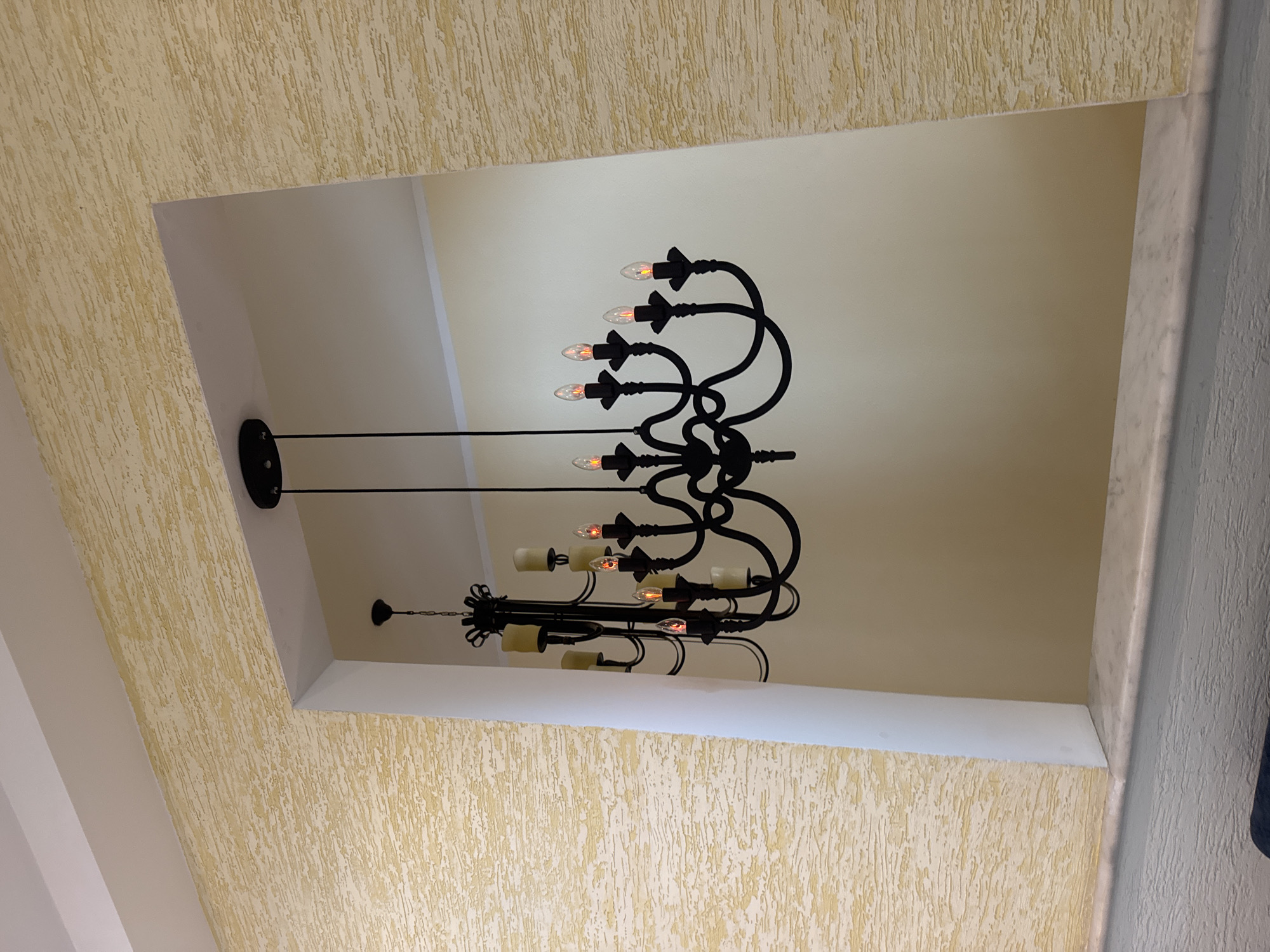 A low-angle shot shows a black metal chandelier hanging within a recessed ceiling structure. The ceiling is a light, creamy color, contrasting with the dark metal of the chandelier. The chandelier has multiple arms, each ending with a light fixture that resembles a candle. Some of the "candles" are lit, casting a warm glow. The recessed area is framed by textured, yellow walls that appear to be made of stucco. The right edge of the frame contains a white wall, with a dark blue object just visible along the side. The location is unknown.
