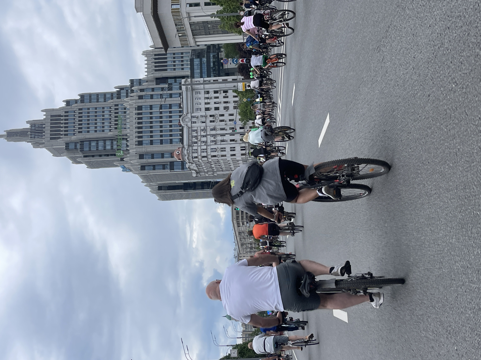 A large group of cyclists is riding on a wide asphalt road in Moscow, Russia. The perspective is from behind the cyclists, looking toward tall buildings. The sky is overcast. In the foreground are two cyclists; one is wearing a white shirt and gray shorts, the other a gray shirt and black shorts. Other cyclists are visible further ahead. In the background are buildings, notably a stepped, modernist building with many windows. Street lights are visible on the left side of the road. White lane markings are present on the road surface. The environment suggests an urban setting.