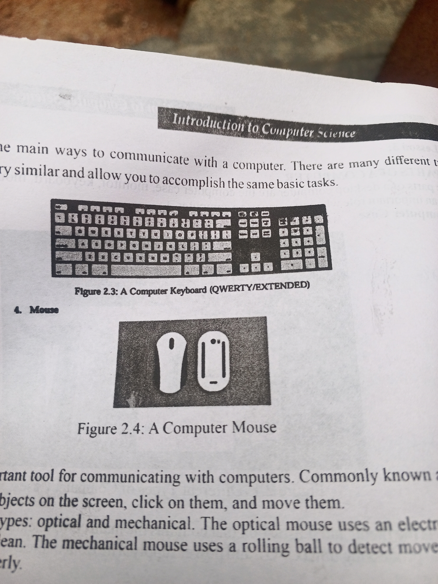 The image is a photograph of a page from a book or document, depicting an introduction to computer science concepts. It features a title "Introduction to Computer Science" and two figures: Figure 2.3, a QWERTY keyboard, and Figure 2.4, a computer mouse. Text accompanying the figures describes their function in communicating with computers. There is no discernible information about Jalingo, Nigeria, or any people present in the image. The environment is that of a flat surface, likely a table, with some textured background elements that are out of focus.