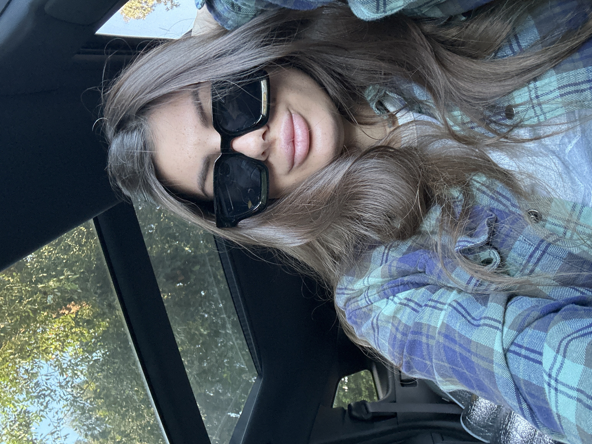 A woman wearing sunglasses and a blue plaid shirt is lying down inside a car, possibly looking up at the sunroof. Outside the car, trees and foliage are visible. The location is Unknown, Unknown.
