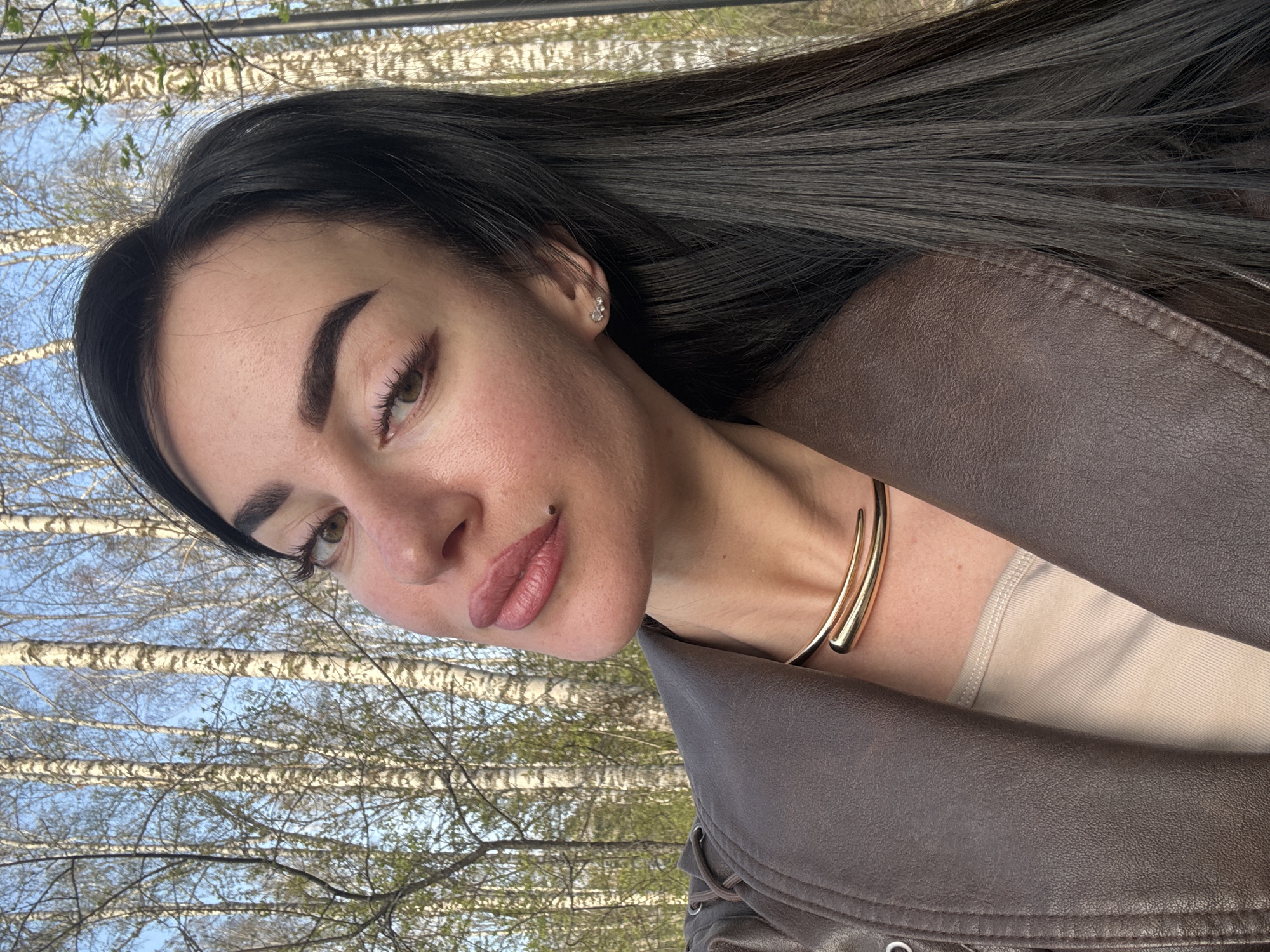 A woman with long, dark hair is positioned in front of a backdrop of birch trees. She is wearing a brown leather jacket over a beige top and a gold-colored double-strand necklace. The photo appears to be taken outdoors, with the trees suggesting a park or wooded area. There is no specific information indicating the location in Krasnoyarsk, Russia.
