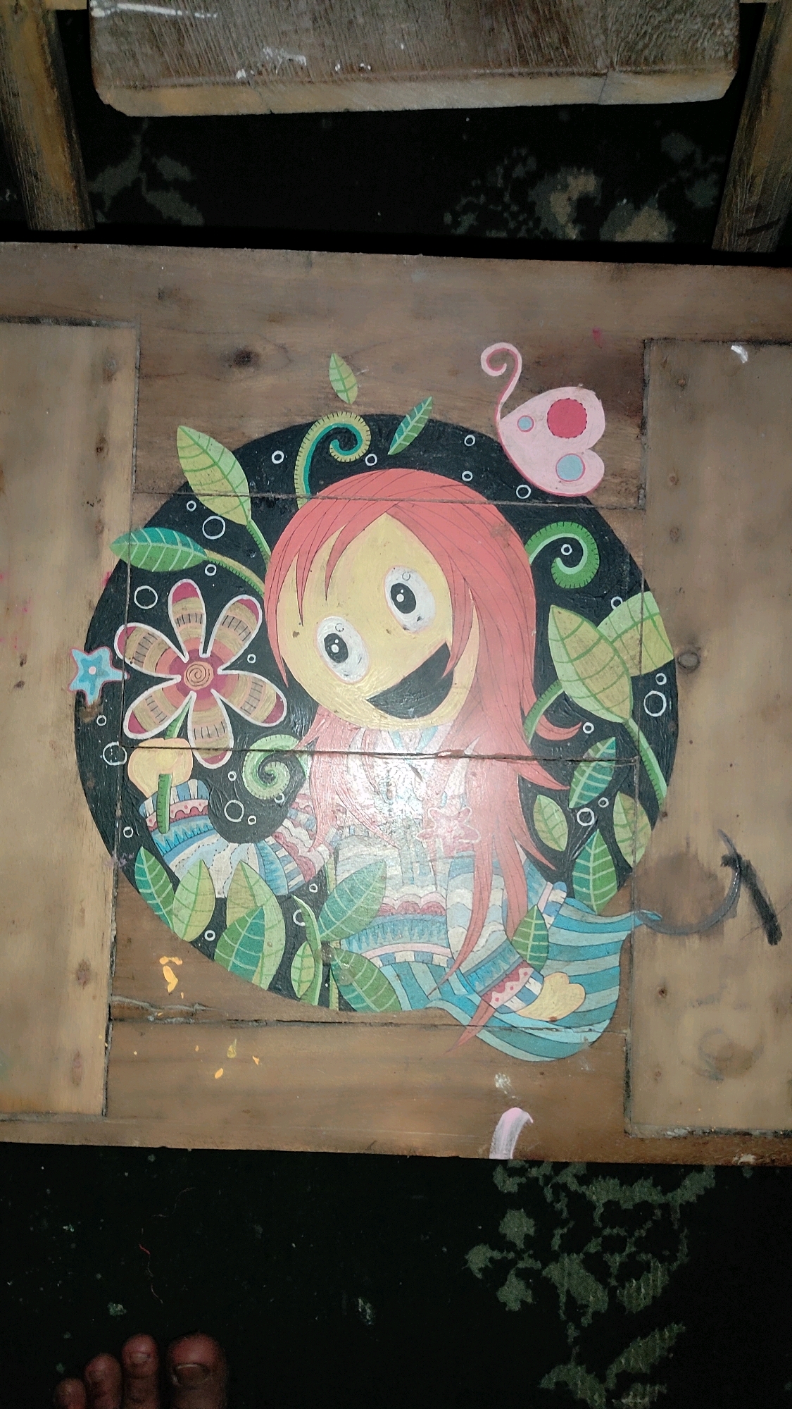A painting on a wooden surface depicts a cartoonish character with red hair, wide eyes, and an open mouth, surrounded by leaves, flowers, and a pink butterfly. The painting appears to be on a table or bench outdoors, as indicated by the rough wooden planks and the presence of a foot in the lower left corner of the frame. The background is dark, suggesting it may be night or a shaded area. The style of the painting is whimsical and colorful. The location of this image is Guwosari, Indonesia.