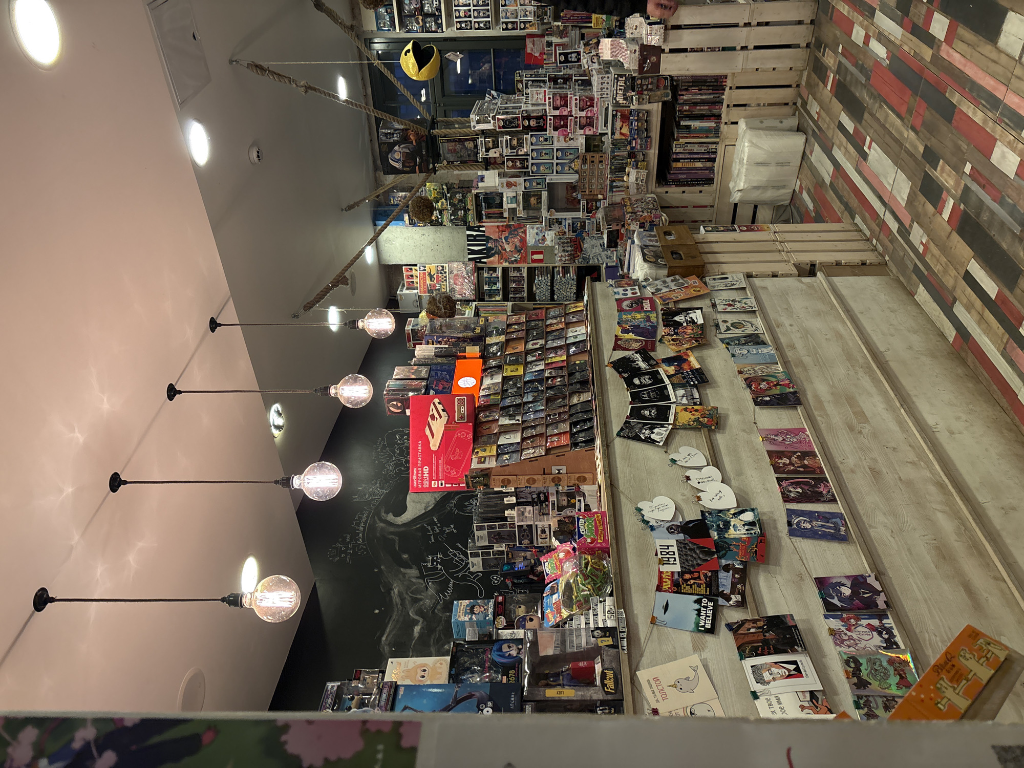 This is an interior shot of a shop in Saint Petersburg, Russia, with a cluttered, maximalist aesthetic. The space is packed with merchandise and displays.
On the left side of the frame are light pink walls with a series of exposed light bulbs hanging from long cords. Adjacent to the wall is a black chalkboard covered in mathematical equations, drawings, and handwritten notes. On the right side, a wall is clad in multi-colored wooden planks, and a makeshift staircase of wooden crates leads up to shelves filled with DVDs and collectibles.
The shop floor is covered with books, comics, games, and other merchandise. A large red box with the words "ИГРОВАЯ ПРИСТАВКА BitHD" in Cyrillic text is prominently displayed. Other items include a yellow Pac-Man figure and numerous Funko Pop figures. The items are a mix of pop culture and local interest products.
In the foreground is a partial view of a low display case with additional merchandise.