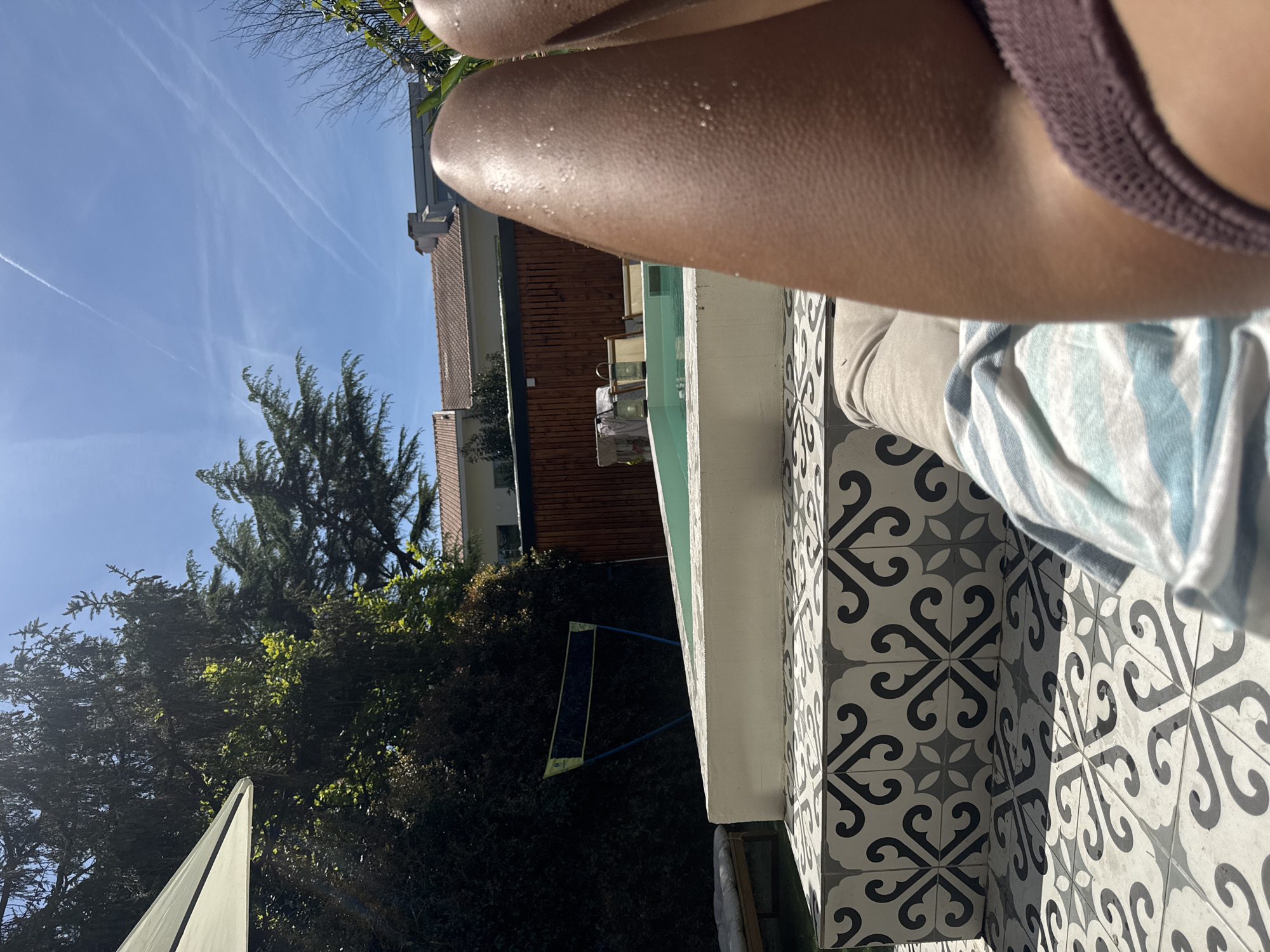 A first-person, upward-angled, eye-level shot reveals a person seated by a pool in an outdoor setting. The individual wears a mauve crocheted top and beige trousers. A blue-and-white striped towel rests in their lap. The pool’s edge is tiled with black and white decorative tiles. The pool water is a light green color. Beyond the pool, a wooden-clad building with a terra cotta roof can be seen. Lush greenery and trees fill the background, extending towards a clear blue sky. A volleyball net is also visible in the background. The presence of European architecture and flora suggests a location in Porto, Portugal. The scene conveys a relaxing, sunny atmosphere.