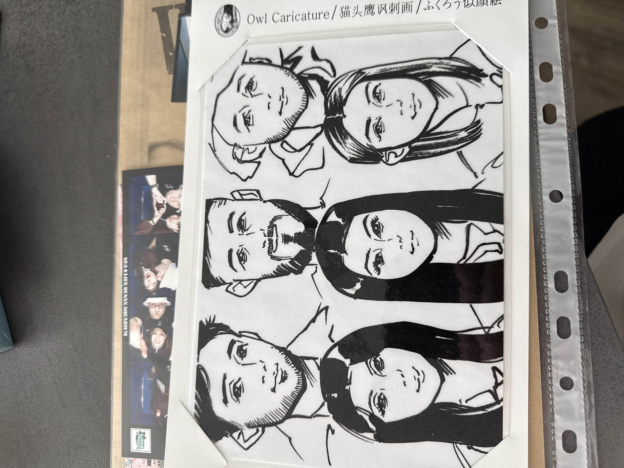 The image shows a drawing labeled "Owl Caricature" in English, Chinese, and Japanese. The drawing features three pairs of portraits in a cartoonish style, likely caricatures of individuals. The top pair is sketched with thinner lines, while the two pairs below have progressively thicker and bolder lines, with larger areas filled in black. The print is enclosed in a white frame, and partially obscured by a clear plastic sleeve or binder sheet with punched holes. To the left of the framed drawing is a vertical strip of photos, possibly a photo booth strip, depicting people. The text "SEA LIFE IRYSAN AQUARIUM" can be partially read on it. The objects rest on a dark surface. The location is unknown.