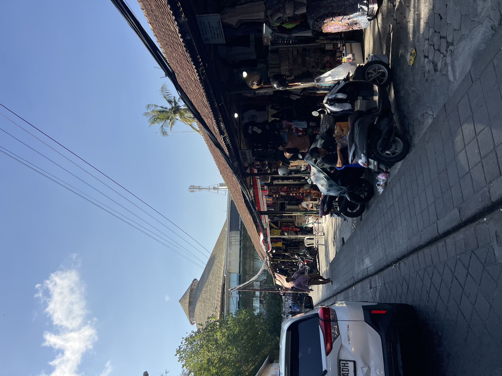 A street scene in Kuta, Indonesia, is depicted. On the right, a row of market stalls with brown tiled roofs lines the street, displaying various goods such as clothing and accessories. Several scooters are parked along the sidewalk in front of the stalls. Two men are seated within the stalls. On the left, a white SUV is parked, and a man is walking down the street. In the background, a large building with a thatched, conical roof is visible, along with trees and a communication tower. The sky is clear and blue with some scattered clouds. The street is paved with cobblestones.