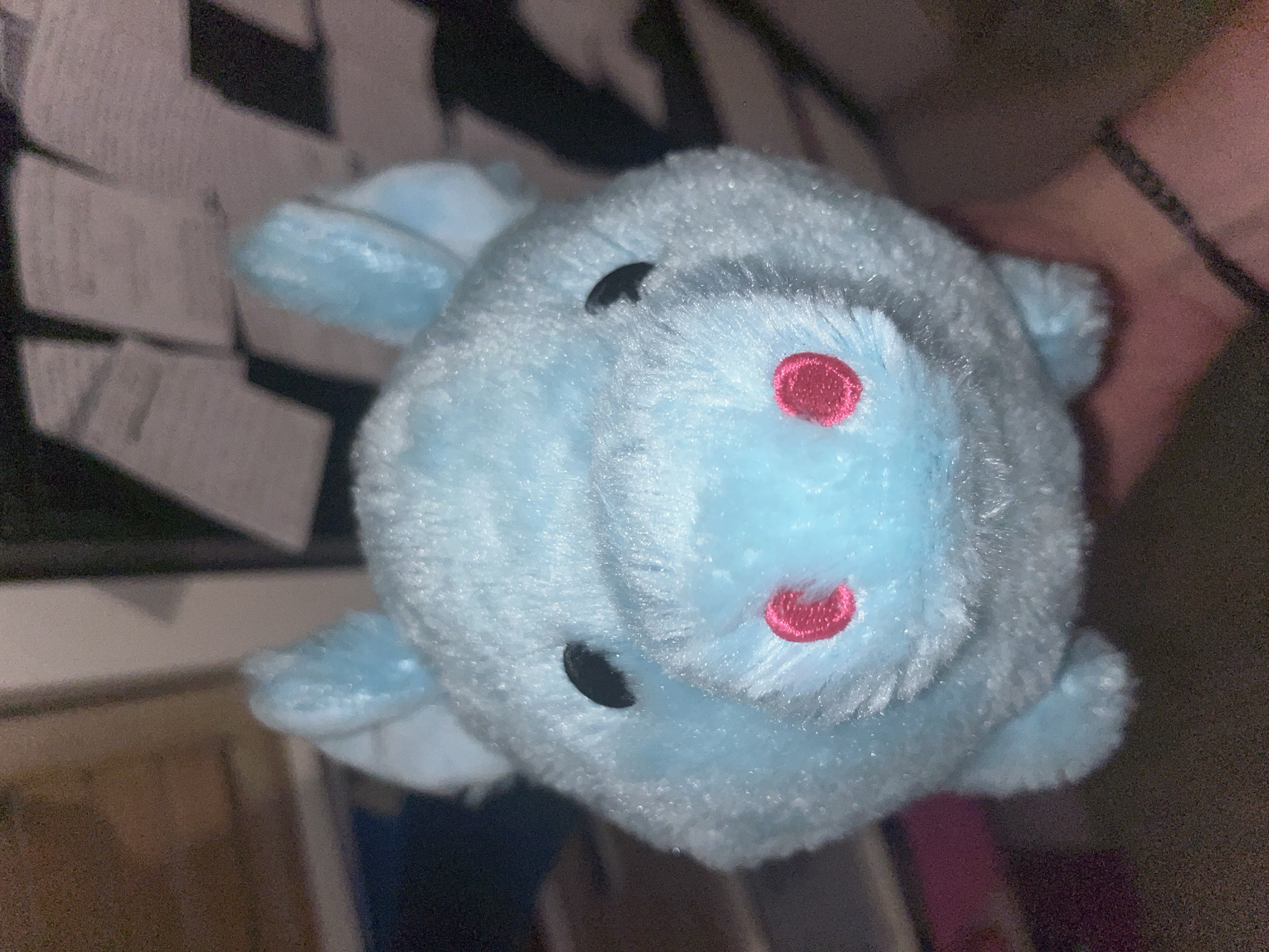 This media file is a close-up, upside-down, and slightly blurry image of a plush toy. The toy is light blue and appears to be some kind of animal, possibly a pig or a bunny, with two black eyes and a pink embroidered nose. Its fur is fluffy and textured. The background is out of focus and dark, with some lighter rectangular shapes visible, suggesting it might be a wall with papers or posters. A hand, with a bracelet, is holding the toy from the upper right side of the frame. The lighting is dim, creating some glare on the toy's fur. No text is visible. The overall impression is that of a casual, perhaps domestic, setting.