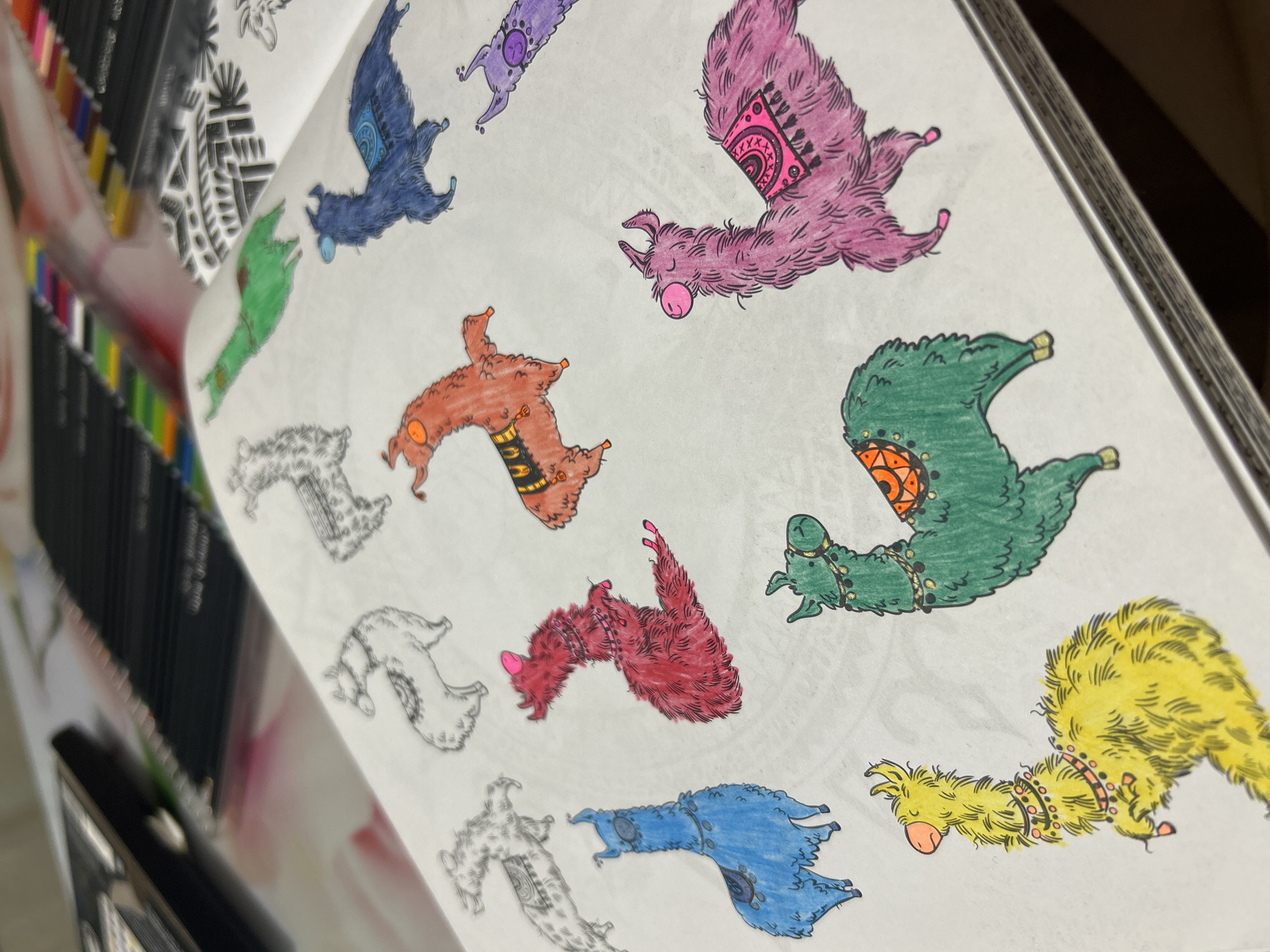 The image displays an open coloring book featuring multiple stylized llama or alpaca creatures. The perspective is an angled, close-up shot looking down into the book.
On the white pages of the book, several llama-like figures are visible. Many are fully colored, exhibiting vibrant hues such as purple, magenta, dark blue, orange-brown, deep red, forest green, and bright yellow. Each colored creature is depicted with detailed patterns on their backs, resembling saddles or blankets, and some also have collars or small bells. The coloring style appears to be done with markers or fine-tipped pens, giving a distinct texture to the "fur" of the animals. In contrast, other creatures on the page are left as black outlines or partially colored, indicating an ongoing activity.
To the left of the coloring book, a collection of colored markers or pencils in a black case is partially visible and out of focus, suggesting the tools being used for the activity. The scene takes place indoors, likely on a flat surface such as a table or desk, as someone is engaged in coloring. No specific time of day or weather conditions can be determined, but the activity itself implies a calm and creative environment. There is no visible text in the image.
