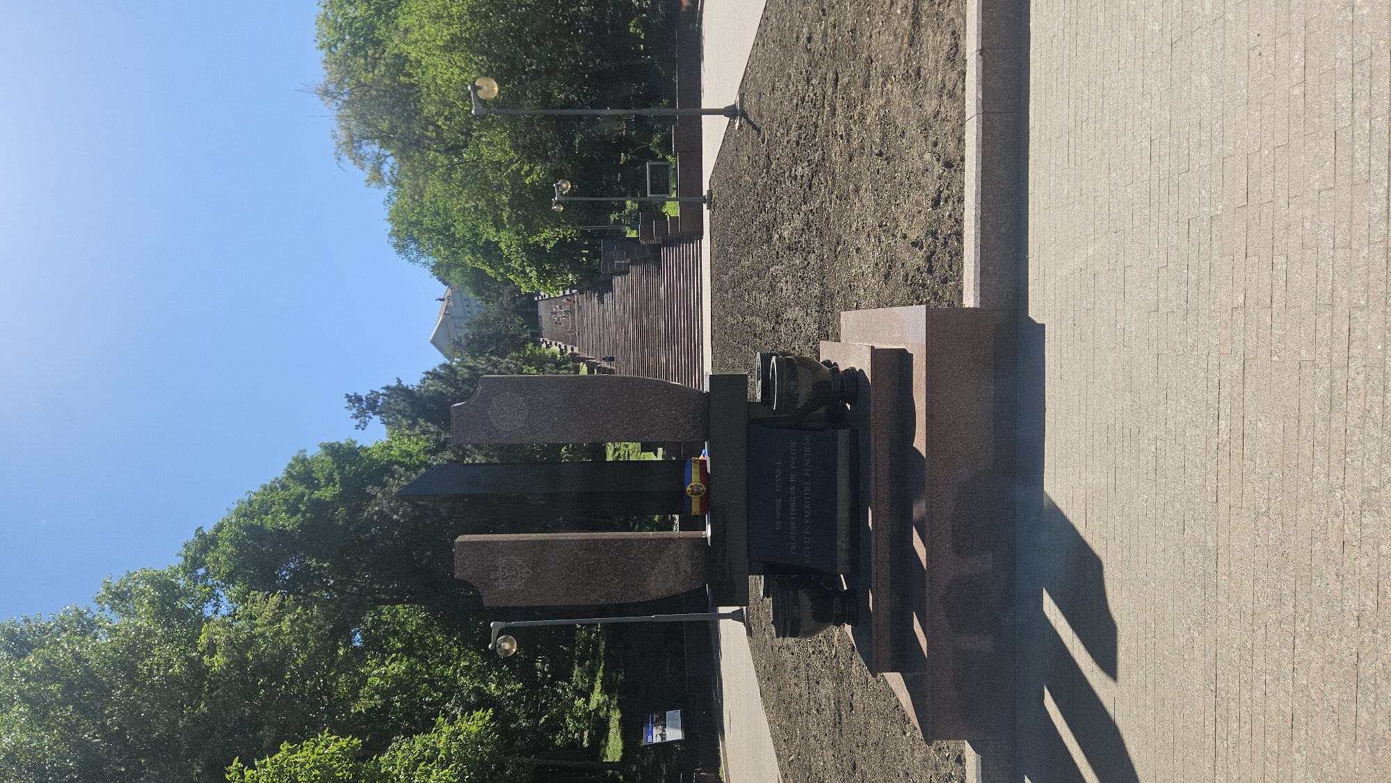The image depicts the "Monument to the Victims of Soviet Occupation" in Chișinău, Moldova. The monument is composed of granite slabs and features sculpted elements. A plaque with an inscription is visible. The monument is situated near a staircase leading uphill through a park with mature trees. Street lamps are placed along the pathway. The day appears sunny and clear. Paved walkways flank the monument.