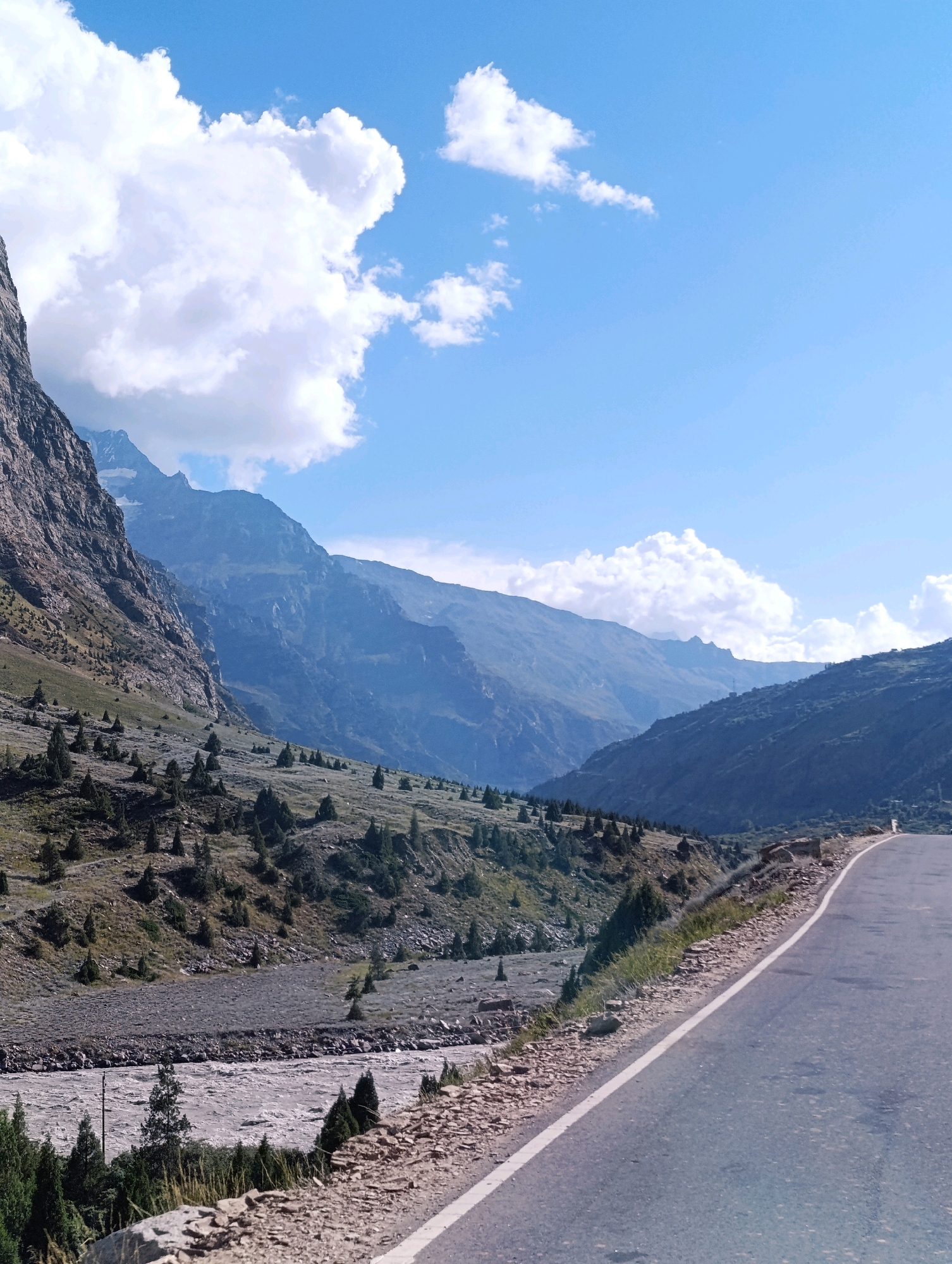 The image depicts a scenic mountain landscape with a road in the foreground. The road curves to the right, leading into a valley. On the left side of the road, a steep, rocky mountain slope is visible, dotted with sparse vegetation and small trees. A wide, shallow river flows through the valley below the slope. In the background, several layers of mountains recede into the distance, with the furthest peaks appearing hazy and blue. The sky is a clear blue, with a few scattered white clouds. The location is identified as Gemur, India. There are no people or animals visible in the image. The scene is a natural landscape, likely a highway or road passing through a mountainous region.