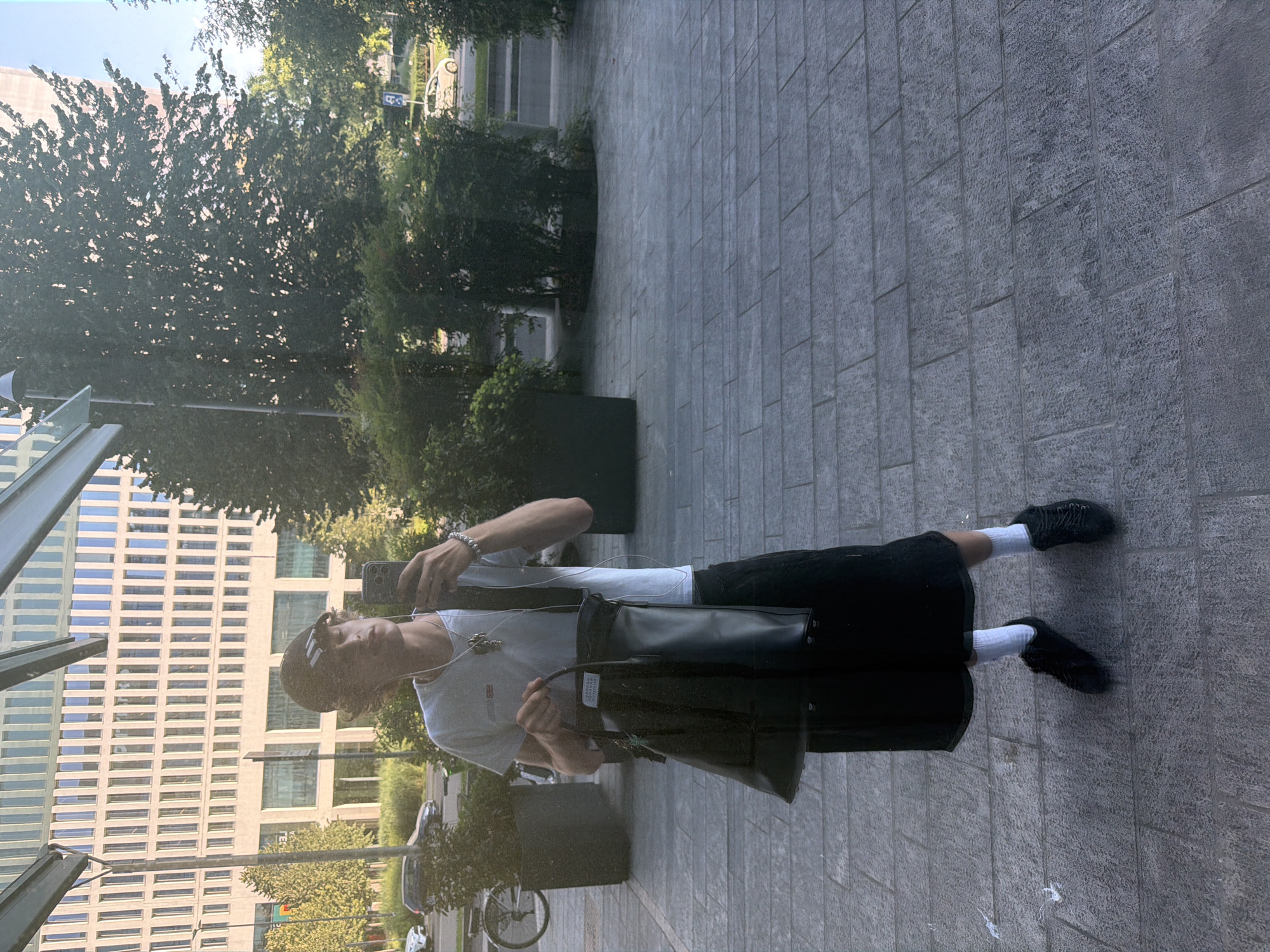 The image shows a young person in a white t-shirt and black skirt taking a selfie with their phone. They are wearing a black baseball cap and holding a black handbag. The person is standing on a paved walkway in front of a modern building with a glass facade. Trees and bushes are visible in the background, along with a street and a few cars. The overall impression is that of an urban environment, possibly in Milan, Italy, given the architectural style of the building and the presence of lush greenery. The lighting suggests it is daytime, with sunlight filtering through the trees.