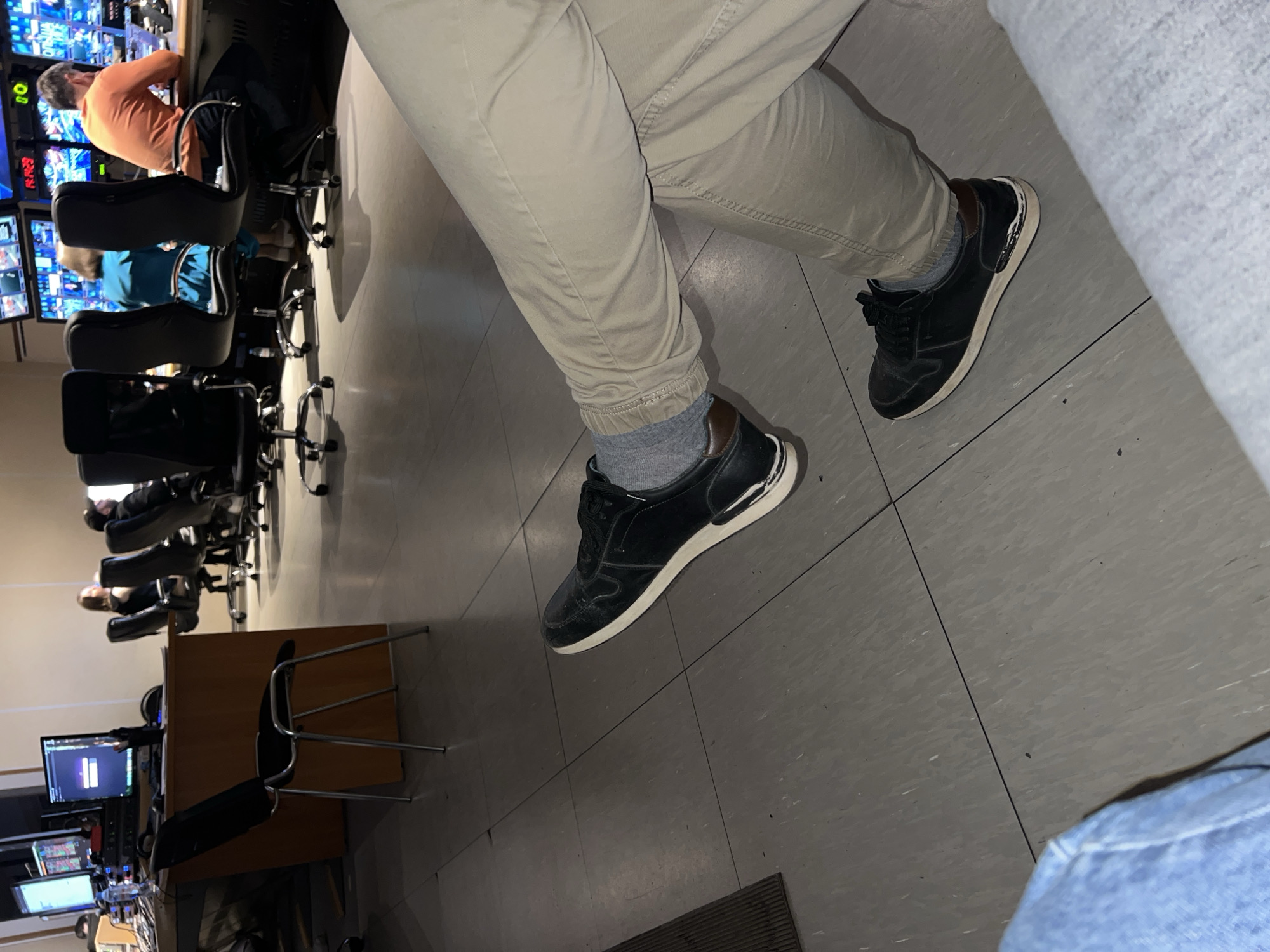 The image depicts an indoor scene, likely a control room or media production studio in Khimki, Russia. In the foreground, the lower half of a person's body is visible, wearing light-colored (khaki or tan) jogger-style pants, grey socks, and black casual sneakers with white soles. This person appears to be seated or standing close to the camera, possibly the photographer.
In the background, the room is equipped with multiple black office chairs, some with wheels, and desks arranged in rows. Several computer monitors are prominent, displaying various interfaces that suggest live video feeds, data monitoring, or control panels. One monitor in the far background shows what resembles a login screen, though specific text is unreadable.
Two other individuals are visible. One person, wearing an orange long-sleeved shirt, is seated at a desk, facing a bank of monitors and appearing engaged in work. Another person, wearing a blue shirt, is partially visible further down the row of desks. The overall activity suggests a work environment focused on observation, monitoring, or production.
The lighting is artificial, with the bright screens illuminating parts of the room. The floor is a neutral grey, appearing to be tiled or linoleum. The scene conveys a professional and functional setting. No specific brand names or detailed text are clearly legible within the image.