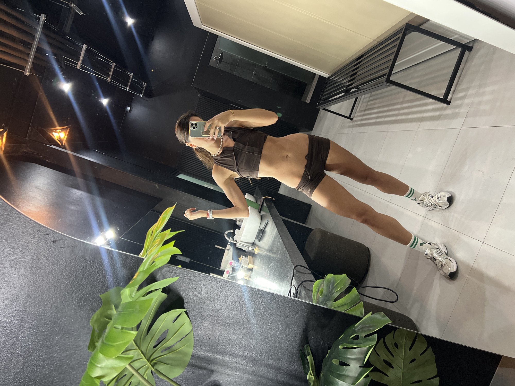 A person is standing in a bathroom and taking a mirror selfie. The person is wearing a brown crop top and brown shorts, white sneakers, and green and white striped socks. They are also wearing a pearl necklace and a bracelet. In the background, there is a black wall, a large plant, a sink, a shower, and a bench. The room appears to be a locker room or gym bathroom.