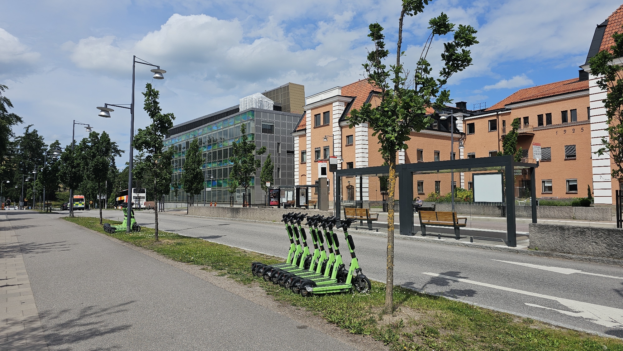 This is a street-level, eye-level, wide-angle shot of a street in Linköping, Sweden. The photo captures a row of bright green electric scooters parked on a grassy verge alongside a paved sidewalk. Across the street, a modern glass-fronted building stands next to an older, orange brick building with the year "1925" visible on its facade. A bus shelter with benches is situated in front of the brick building. The sky is partly cloudy, and trees line the street, casting shadows on the pavement. There are a few people visible, including one person sitting at the bus shelter and another in the distance walking. A bus is also present on the road. The overall scene depicts a typical urban environment with public transportation and shared mobility options.