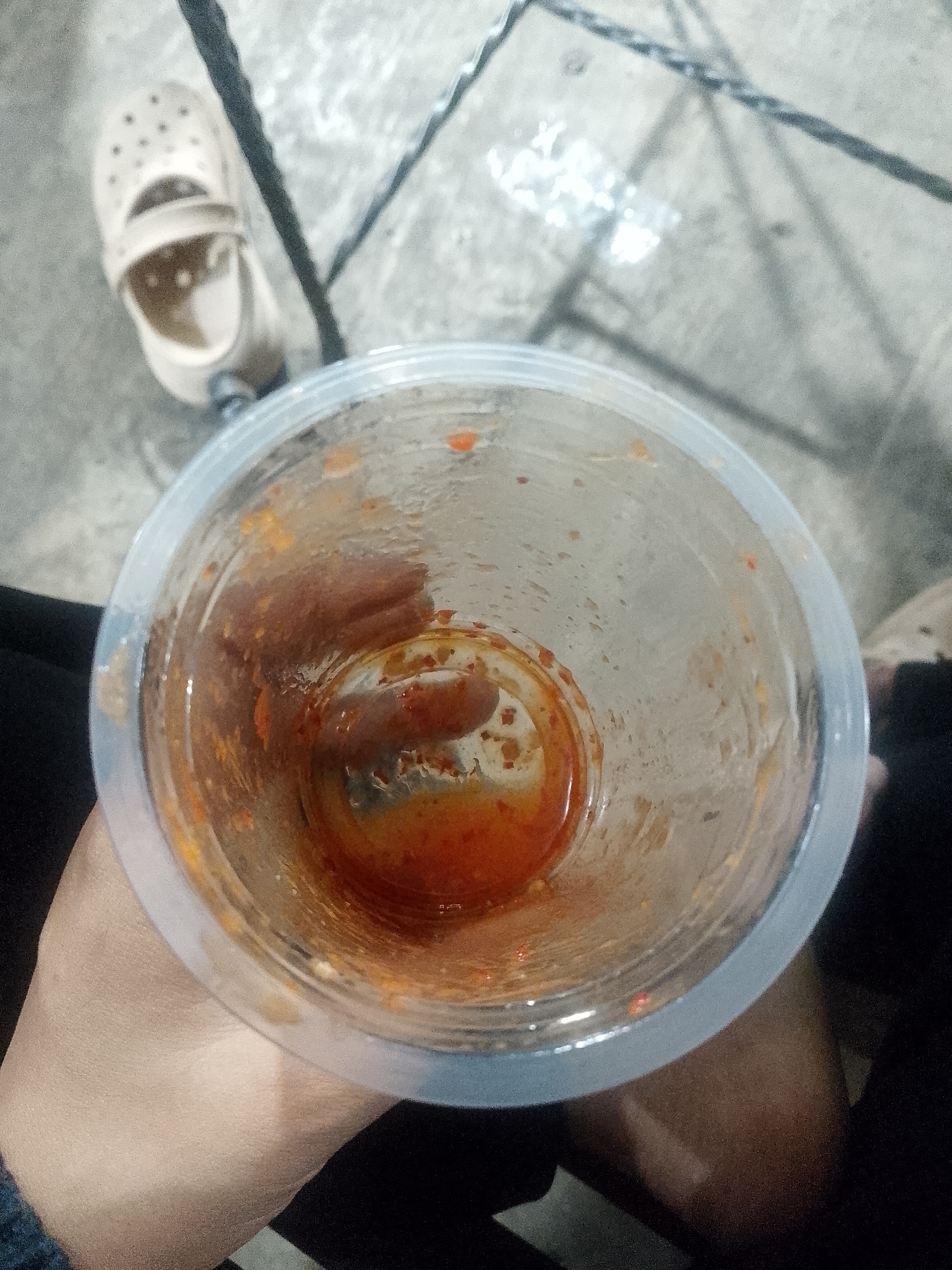 The image is a top-down, close-up shot of a clear plastic cup containing a reddish-orange liquid and some solid remnants, likely food residue. The cup is held by a hand, with a portion of a person's arm and dark clothing visible. The background appears to be an outdoor setting with a textured surface, possibly concrete or tile, and dark linear elements that could be shadows or part of a structure. A light-colored shoe, resembling a clog, is visible in the upper left portion of the image, suggesting a casual or outdoor environment. The lighting is dim, creating some glare on the cup. The scene does not contain any specific landmarks or visual cues to definitively place it in Lhokseumawe, Indonesia.