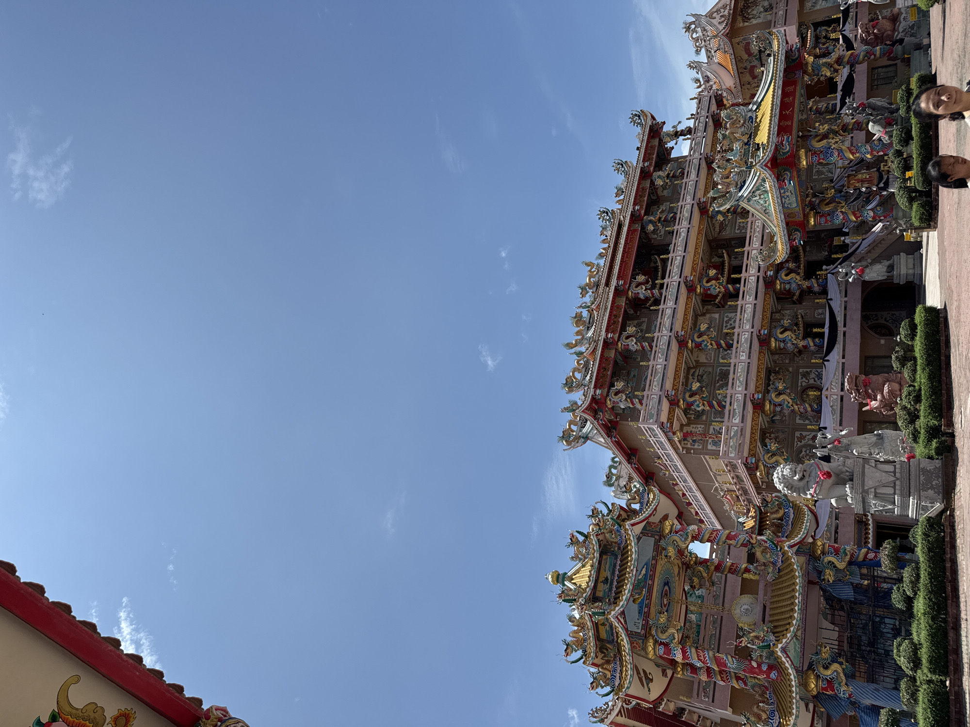 In Ang Sila, Thailand, a vibrant, multi-tiered temple stands against a clear blue sky. The temple's architecture is highly ornate, featuring intricate dragon motifs, colorful sculptures, and tiered roofs adorned with gold and red trim. Two stone lion statues are positioned in front of the temple, one white and one reddish-brown, with red ribbons tied around their necks. Lush green bushes line the foreground, and a paved area in front of the temple is visible. In the lower right corner, two women are present, their faces partially visible. The temple appears to be a significant religious or cultural landmark.