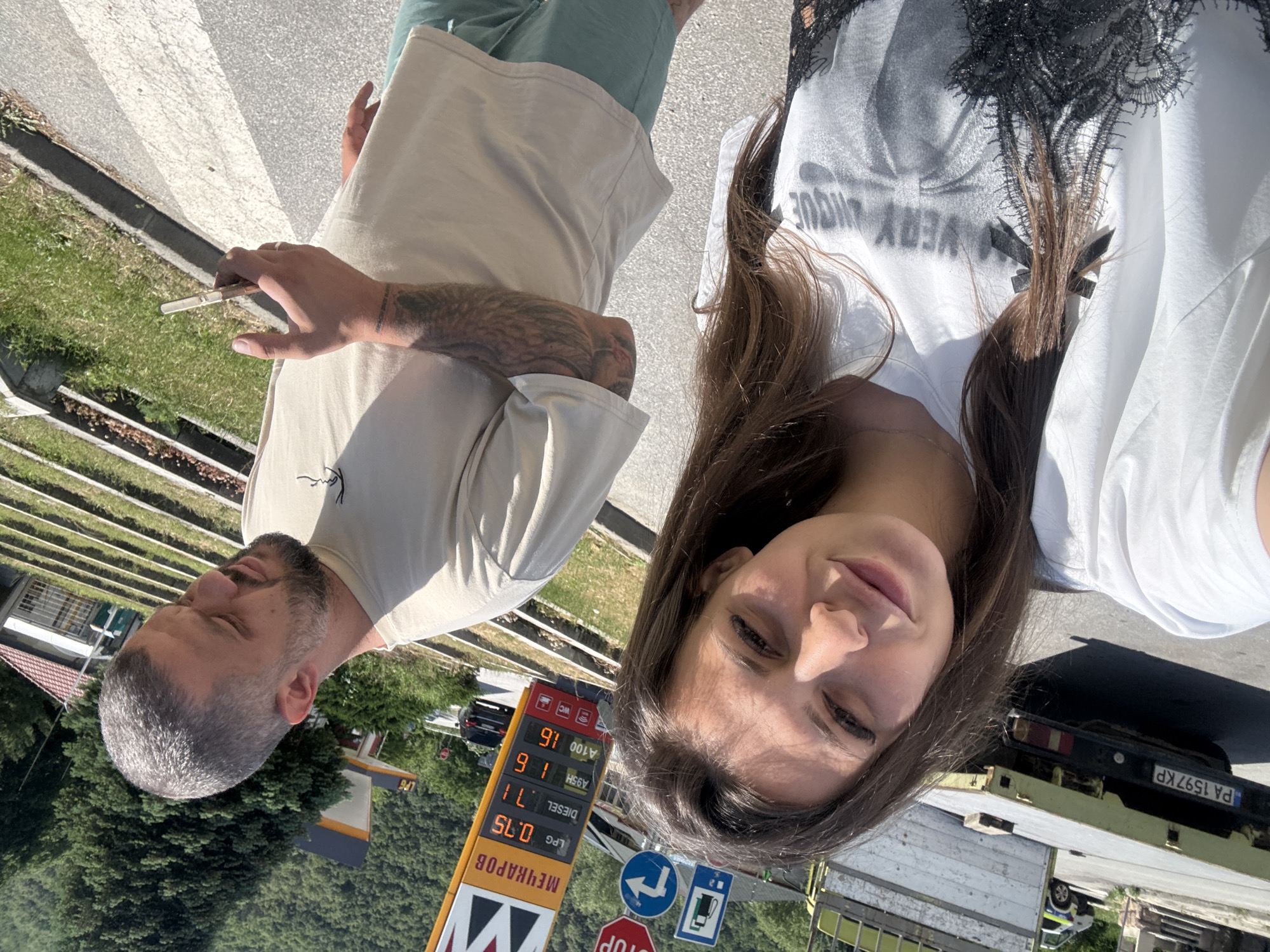 A bright, sunny upward-angled selfie captures a man and a woman at a roadside location, likely a gas station in Vetren Dol, Bulgaria. The image, taken in daylight with strong shadows, shows the subjects from the chest or waist up.
The man, positioned on the left, has close-cropped grey hair and a beard. He wears a light-colored t-shirt and green bottoms. Tattoos adorn his right forearm. He is holding what appears to be a hand-rolled cigarette or joint between his fingers, with his eyes closed in a relaxed expression.
The woman, on the right and closer to the camera, has long brown hair. She wears a white t-shirt with black lace detailing on the shoulder/collar, featuring the mirrored text "VERY INQUE" (likely "UNIQUE"). Her eyes are partially closed, possibly squinting in the bright sun, and she displays a subtle, relaxed smile.
The background reveals elements of a road and a gas station. A prominent yellow and red fuel price sign displays "МЕЧКАРОВ" (Mechkarov) at the top, with prices listed: "LPG 0.75", "DIESEL 1.71", "A95H 1.60", and "A100 1.60". Below the sign, a blue circular road sign with a white left-turn arrow, a red octagonal "STOP" sign, and a blue square sign with a fuel pump icon are visible. A black vehicle with the Bulgarian license plate "PA 1597 KP" is partially seen, along with parts of a yellow/green truck. The setting includes an asphalt road with a crosswalk, grassy verges, and a dense evergreen forest in the distance, consistent with the mountainous region of Vetren Dol, Bulgaria. The overall scene suggests a casual, relaxed stop during a journey.