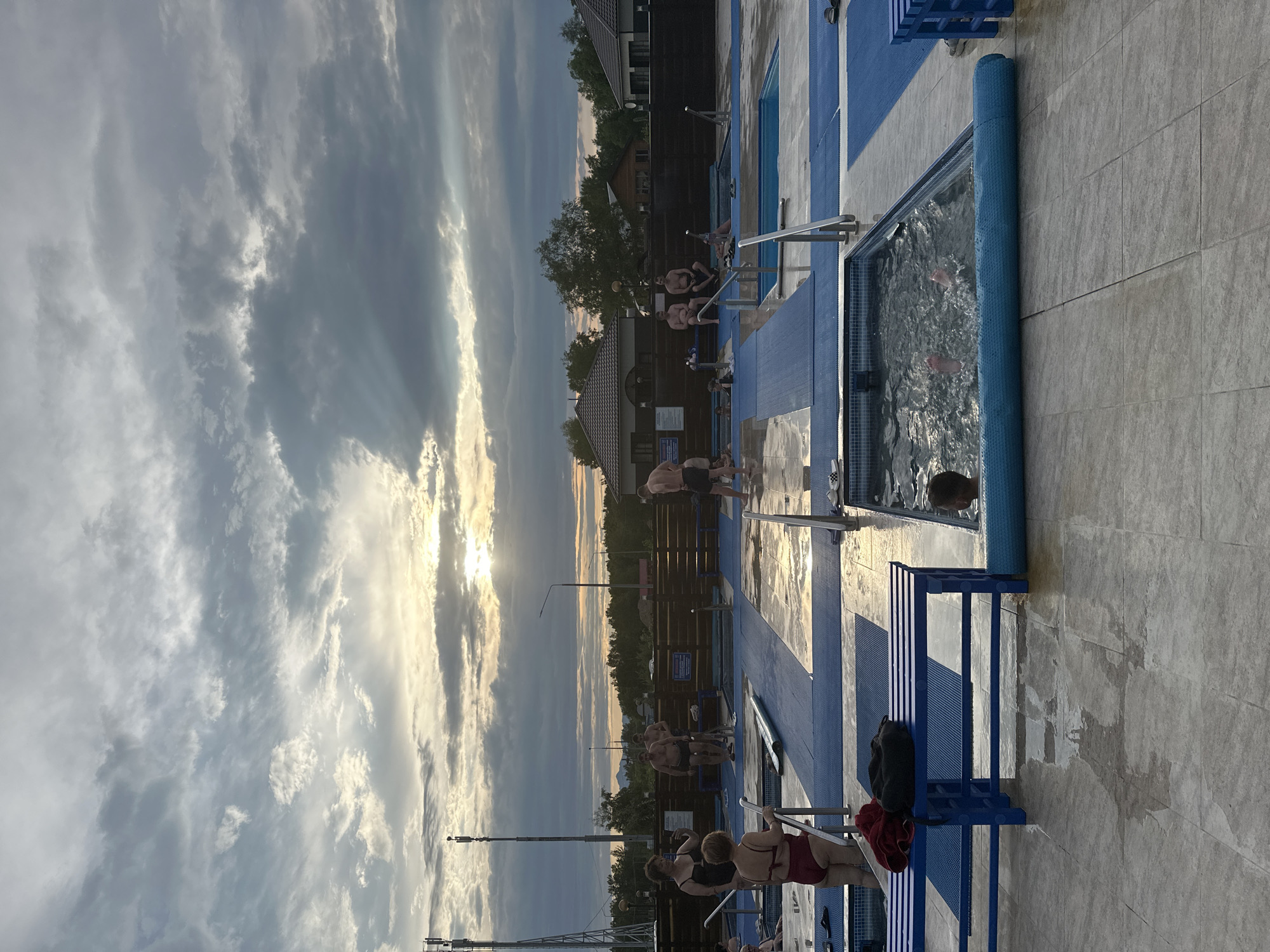 The image depicts an outdoor swimming pool area with several small, tiled pools. The pools are surrounded by blue matting and light-colored tiled flooring, which is wet in places, indicating recent use. Several people are present, some are in the pools, while others are standing or sitting around them. The sky is cloudy with the sun visible through the clouds, casting a warm light. In the background, there are buildings, trees, and telecommunication towers, suggesting an urban or semi-urban setting, possibly in Yuzhnyye Koryaki, Russia.