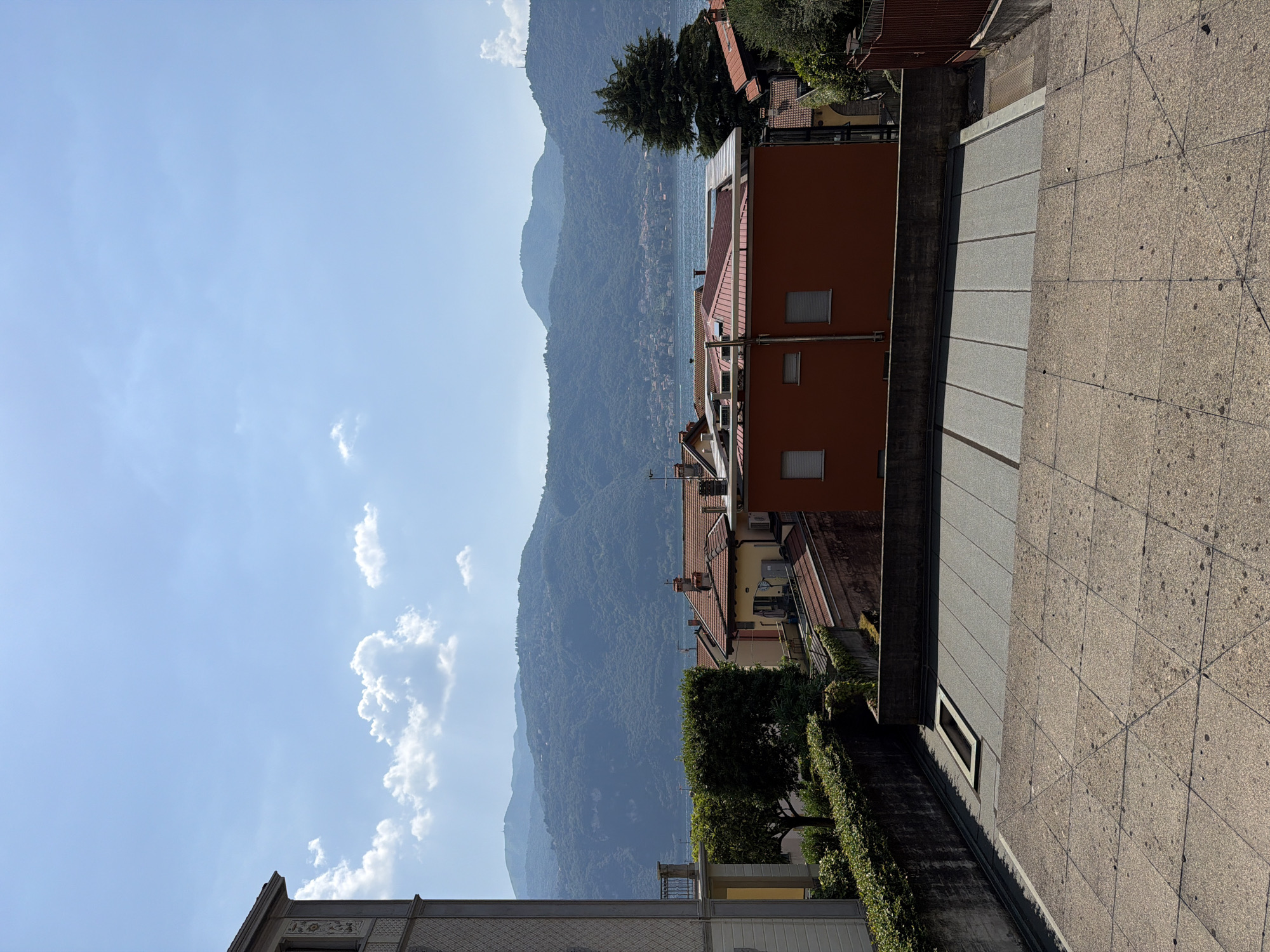 This image captures a serene view from a slightly elevated vantage point in Lierna, Italy. In the foreground, a textured concrete patio with a geometric pattern is visible, leading down a gentle slope. Beyond the patio, a dark gray, flat-topped structure with visible seams and a rectangular opening is present. A concrete wall borders this structure, topped with a dense green hedge.
Further into the scene, the roofs of several buildings are visible. One building, painted a vibrant orange, has a flat roof and two small, white-shuttered windows. Adjacent to it, a building with a yellow facade features a terracotta-tiled roof. Behind these buildings, a body of water, likely Lake Como, stretches across the midground.
The background is dominated by rolling, lush green mountains, their slopes covered in dense vegetation. The sky above is a clear, bright blue, with scattered white cumulus clouds. The overall impression is one of a peaceful, sunny day in a picturesque Italian town overlooking a lake and mountains. No people are present in the image.