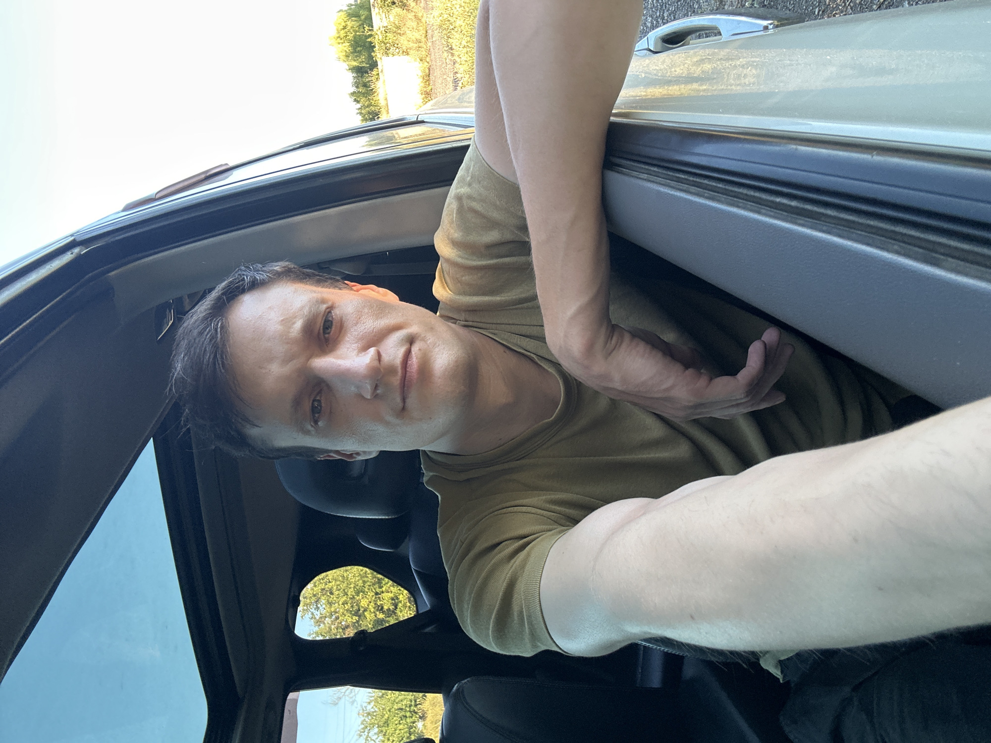 A man is sitting in the driver's seat of a vehicle, with the driver's side door open. He is looking towards the camera, his right arm resting on the open door frame. The man is wearing a light olive-colored t-shirt. The interior of the car appears to be black. A sunroof is visible overhead, letting in natural light. Outside the car, green trees and dry vegetation can be seen under a clear, bright sky. The scene appears to be set in a rural or suburban area, possibly on the outskirts of Makiv, Ukraine.