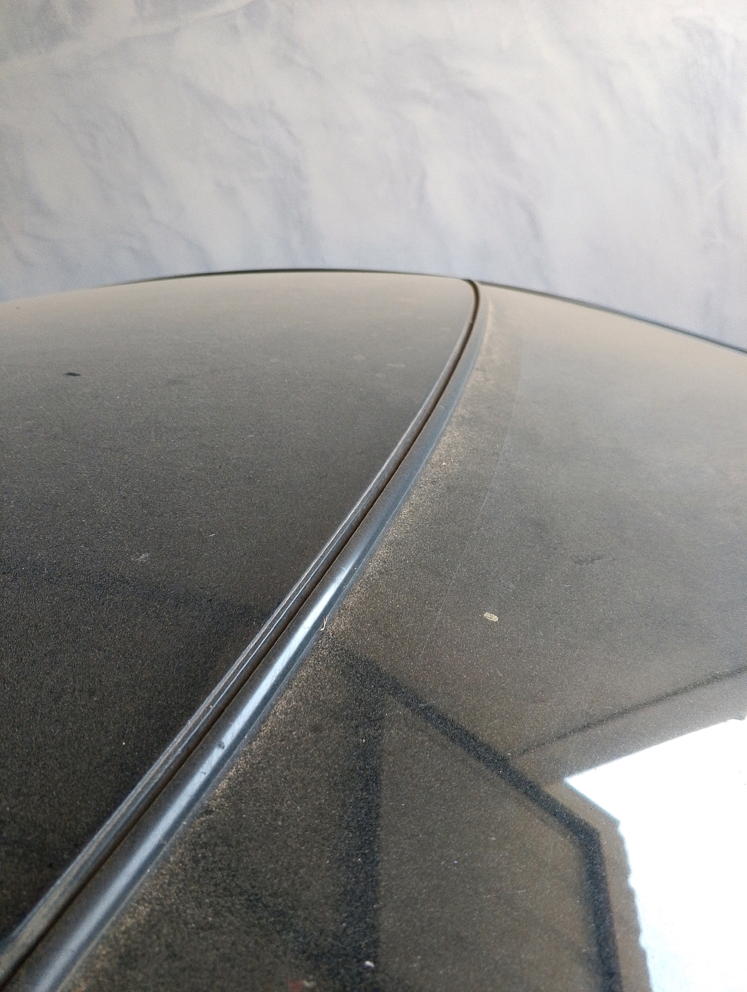 The image shows a close-up, angled view of a dusty car roof. The roof appears to be dark in color, possibly black or dark gray, and it is covered in a fine layer of dust. A prominent seam or trim runs diagonally across the roof from the bottom left to the upper right. The seam is dark and appears to be slightly raised from the surface of the roof. The background of the image is a light, textured surface, possibly a fabric backdrop, that is out of focus. There are no people or other objects clearly visible in the scene. The lighting suggests it is daytime, with a bright reflection visible on the lower right portion of the car roof. The dust on the car suggests it has been parked outdoors for some time.