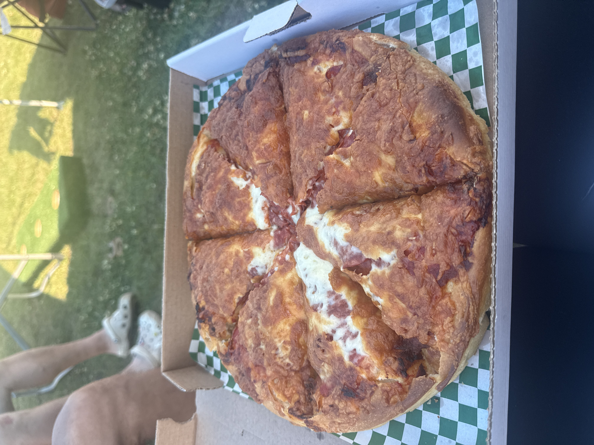 A large pizza, cut into eight slices, sits in a cardboard box lined with green and white checkered paper. The pizza has a thick, golden-brown crust and appears to be topped with cheese and some type of meat. The box is open, and the pizza is the primary subject in the foreground.
In the background, out of focus, are people's legs and feet. One person is wearing white Crocs, and another is wearing patterned Crocs. They are sitting on a grassy area, likely outdoors. To the left of the people, a portion of a cornhole board can be seen on the grass. In the further background, there are shadows cast by what appear to be outdoor chairs and possibly a tent or umbrella. The overall scene suggests a casual outdoor gathering. The context of "city of AB, Canada" is not visually present in the image.