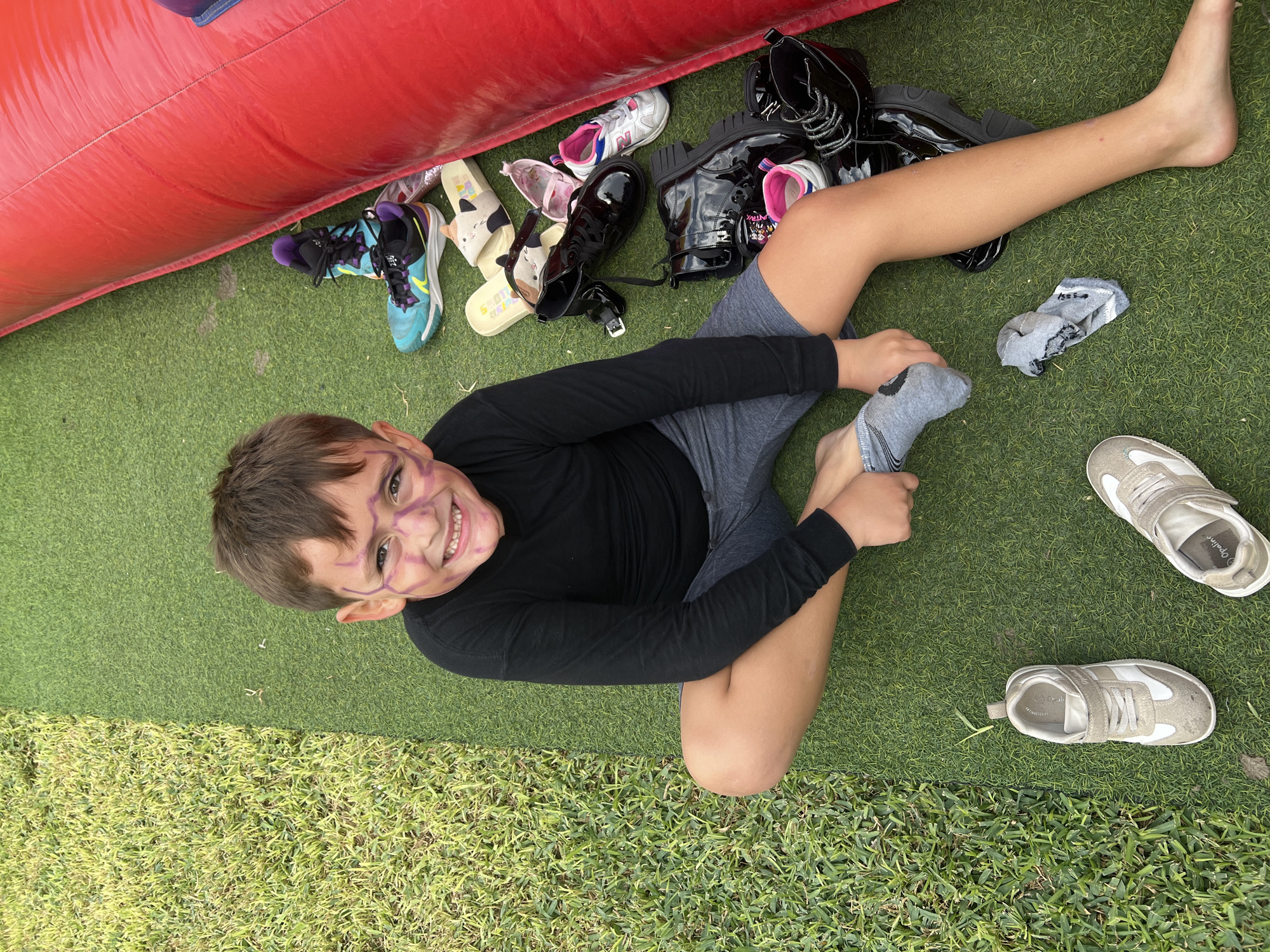 A young boy with brown hair and purple face paint sits on a green artificial turf surface. He wears a black long-sleeved shirt and gray shorts and is putting on a gray sock. Around him are several pairs of shoes, including black boots, sneakers, and sandals. A red inflatable structure is partially visible in the upper left corner. The presence of artificial turf suggests an outdoor recreation area. The location is not discernible based on the information within the image itself. The request to locate this image in the city of Arica, Chile cannot be fulfilled, as the provided image does not contain information to suggest a location.