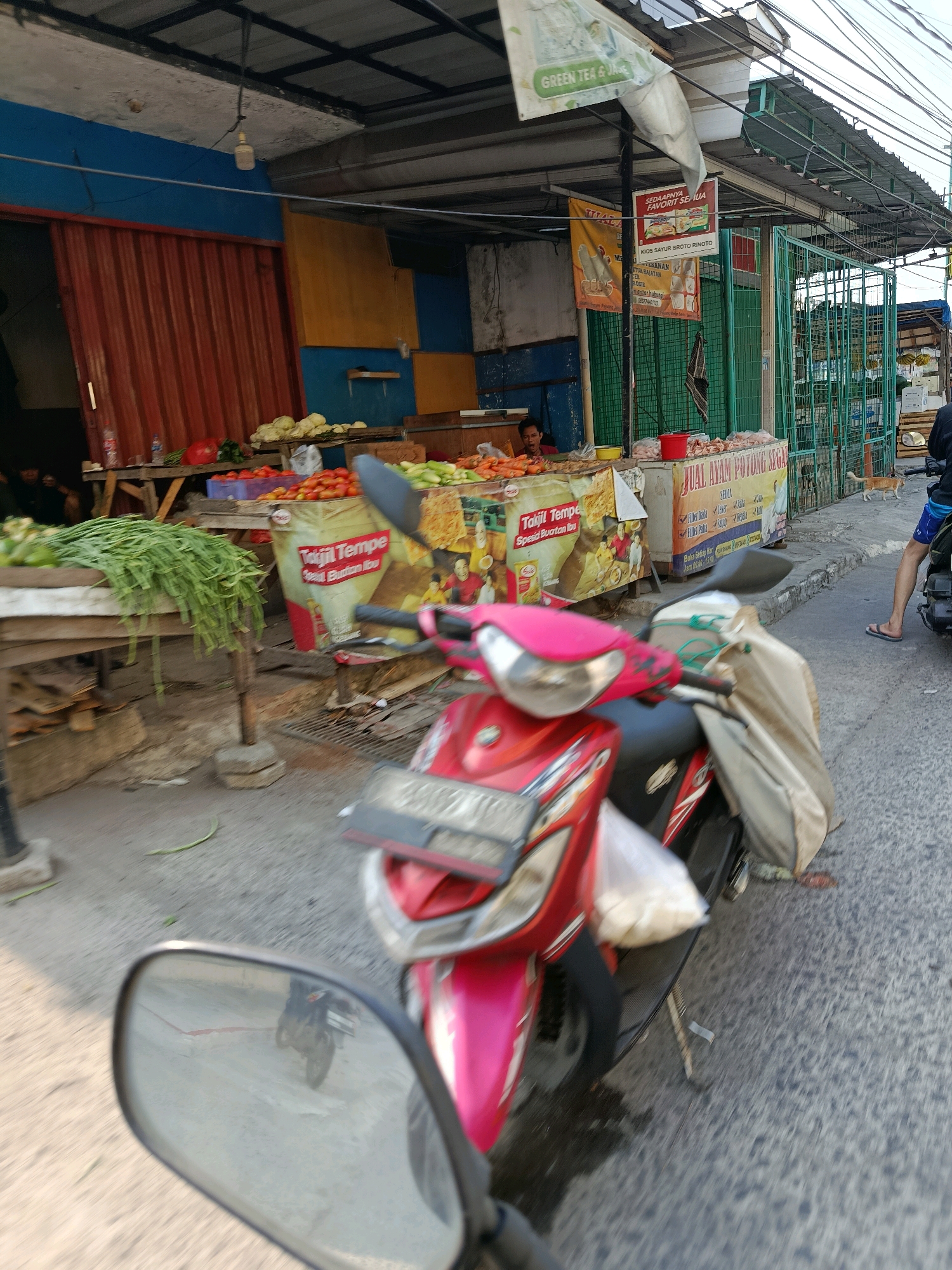 A red scooter is parked on a paved road in front of a market stall. The market stall has a corrugated metal shutter and displays various vegetables, including tomatoes and green beans. There are also posters advertising food products. A person is sitting on the left side of the stall. In the background, a cat walks on the pavement. The scene appears to be in an outdoor market environment in Bekasi, Indonesia.