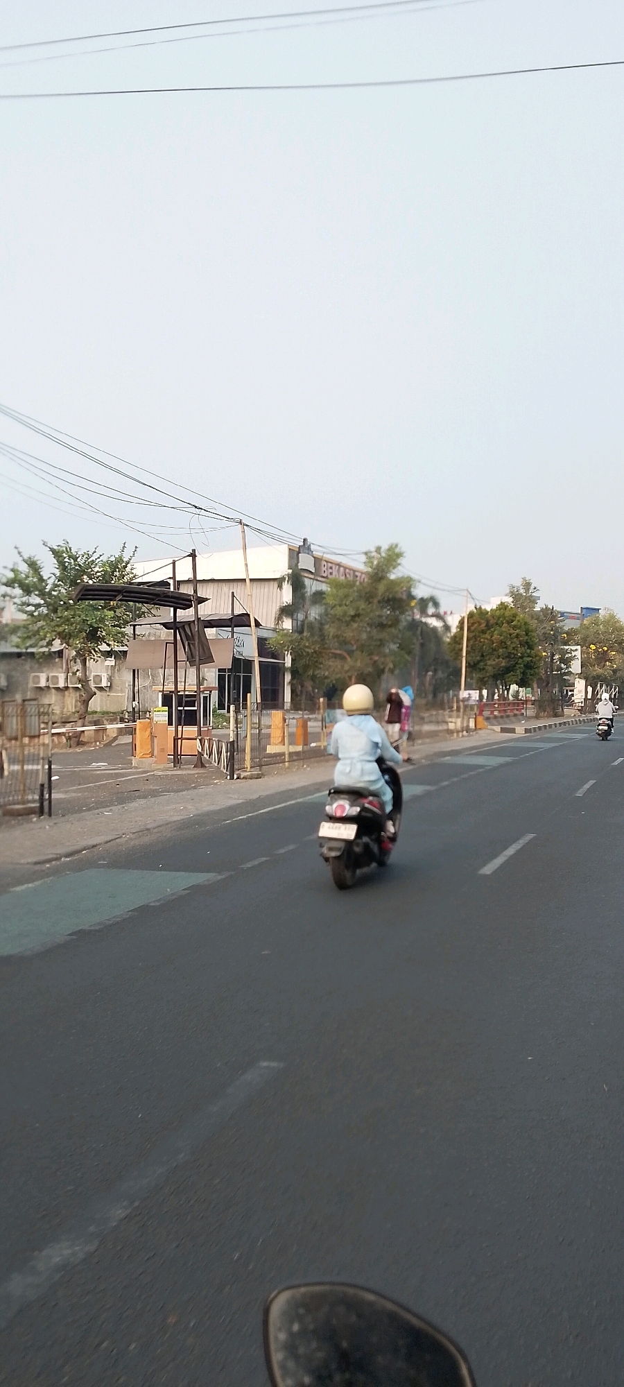The image captures a street scene in Bekasi, Indonesia, likely during daylight with overcast skies. In the foreground, a person is riding a black scooter with a license plate that reads "B 6020 KCK". The rider is wearing a light blue long-sleeved shirt, blue pants, and a light yellow helmet. The scooter is moving along the right lane of a paved road. In the background, there are buildings with signage that appears to say "BEKASI ZONA". Several trees are visible, as are power lines stretching across the frame. There are also metal fences and structures along the side of the road, possibly indicating a business or parking area. Another person in a white hooded jacket is visible riding a scooter further down the road on the right. The overall environment suggests an urban or suburban setting.