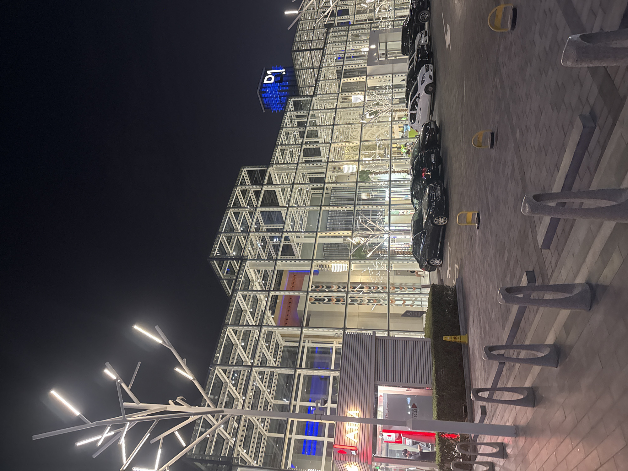The image shows a modern building complex at night, located in Dubai, United Arab Emirates. The architecture features a prominent white metal frame structure with extensive glass paneling. A large blue sign with white lettering "D.1" is visible on the top of one of the buildings. In the foreground, there is a paved area with bollards and a row of parked cars, including black sedans and SUVs, and a white hatchback. A small building with an ATM and a red "ATM" sign is situated to the left of the frame. The scene is illuminated by artificial lights from the building, streetlights resembling stylized trees, and car headlights. The sky is dark, indicating nighttime.