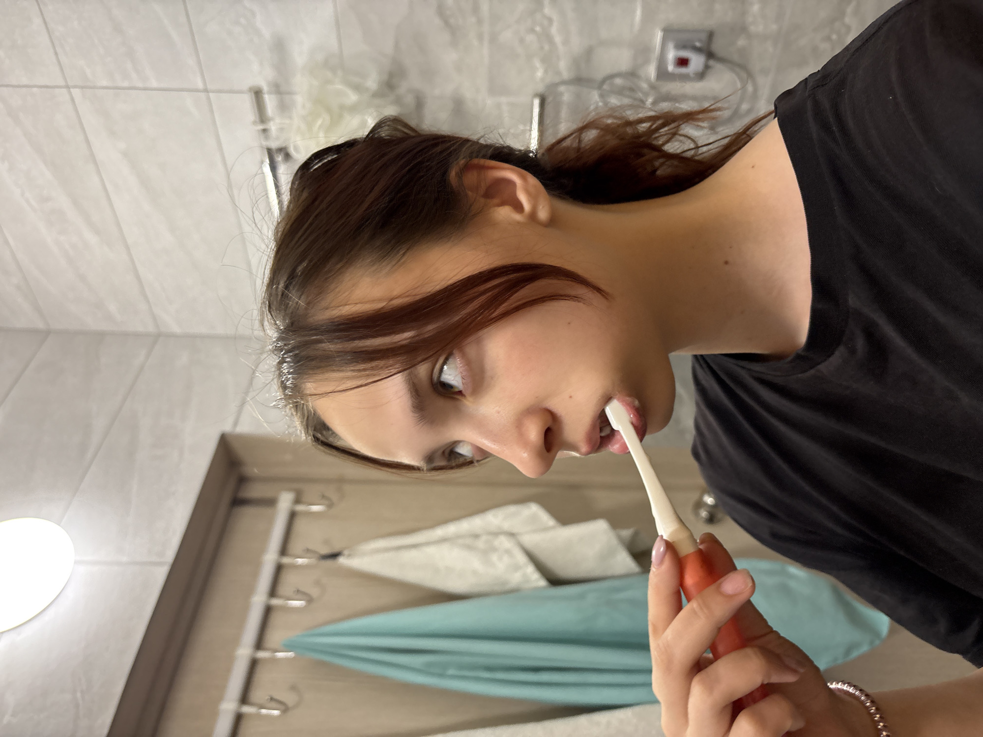 A young woman stands in a bathroom, brushing her teeth. She is fair-skinned with dark hair pulled back, and is wearing a black t-shirt. She is holding a white toothbrush with a red handle to her mouth. The bathroom has light-colored tiled walls and features a light fixture, a towel rack with white and blue towels, and a wall-mounted electrical outlet. The location in the city of Perm, Russia is not discernible from the image.