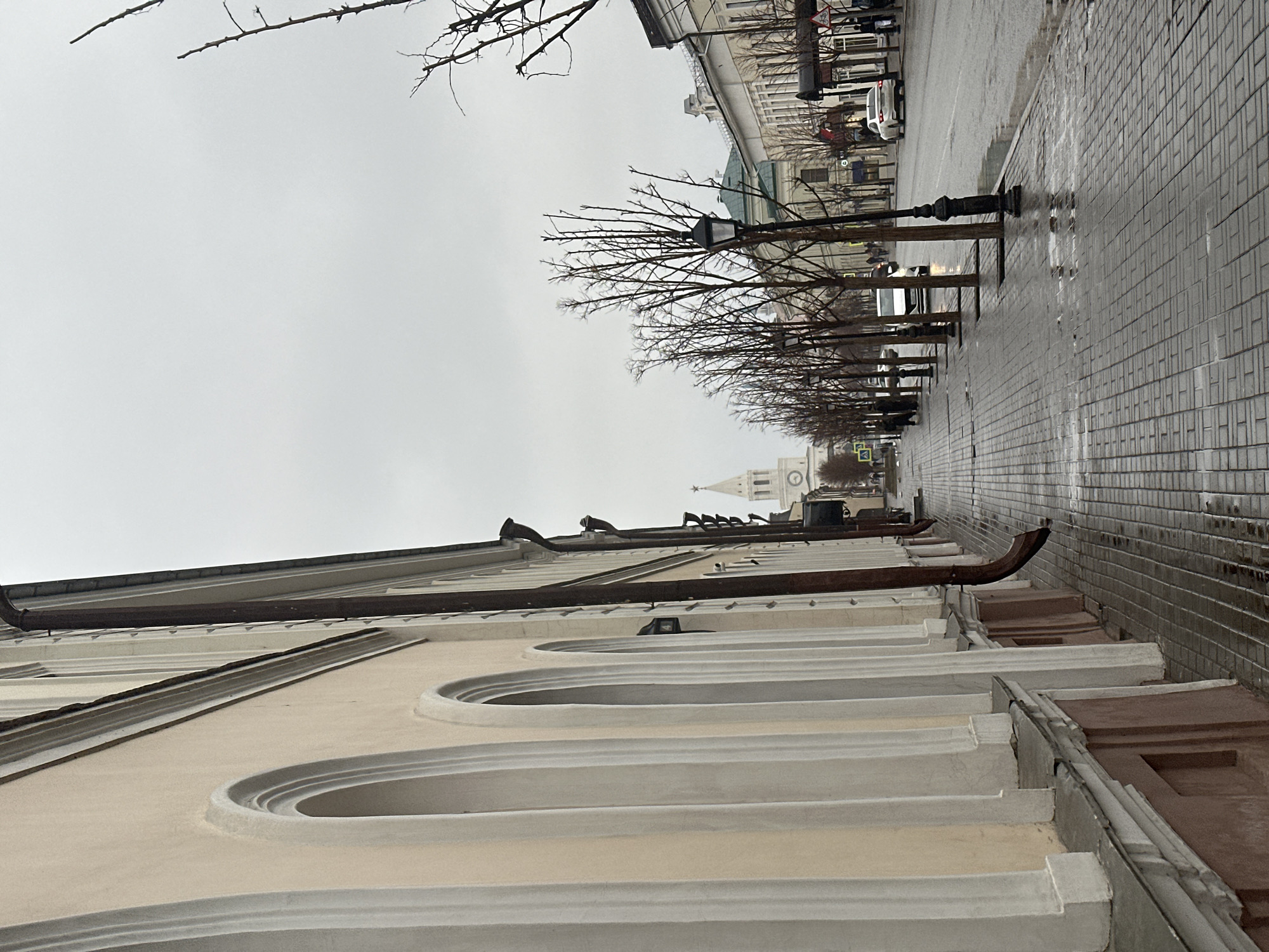 The image captures a street scene in Kazan, Russia. A light-colored building with arched window recesses dominates the foreground. Above the building, a street stretches into the distance, lined with bare trees and old-fashioned streetlights. A tower, likely belonging to a church or historical building, is visible in the distance. The street surface is paved with gray bricks and appears wet, suggesting recent or ongoing rain. Several cars and pedestrians are visible on the street, though they are not sharply defined due to the distance and possibly the weather. The sky is overcast and gray.