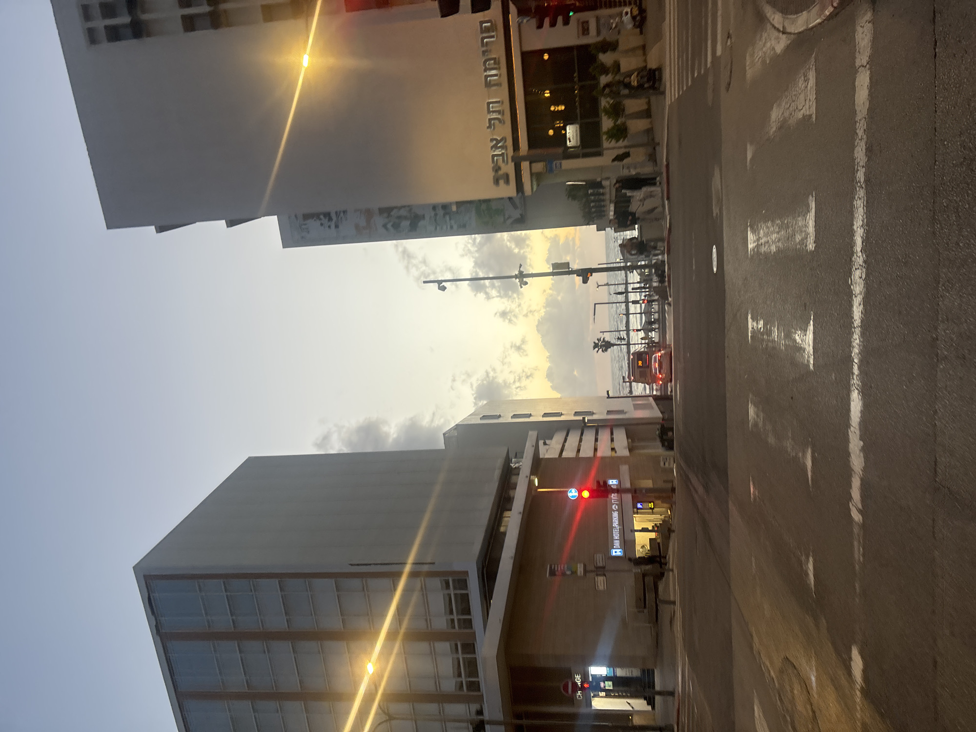 The image shows a street scene in Tel-Aviv, Israel. On the right is a pedestrian crossing with white stripes painted on the asphalt road. The foreground is the road surface. Buildings are visible on both sides of the road. The building on the top has writing in Hebrew, “רמאה תל אביב”. A traffic light is visible in the bottom building, with a red light illuminated. Street lights and traffic lights are also visible. The sky is visible in the middle of the shot. There are no people apparent in the image.