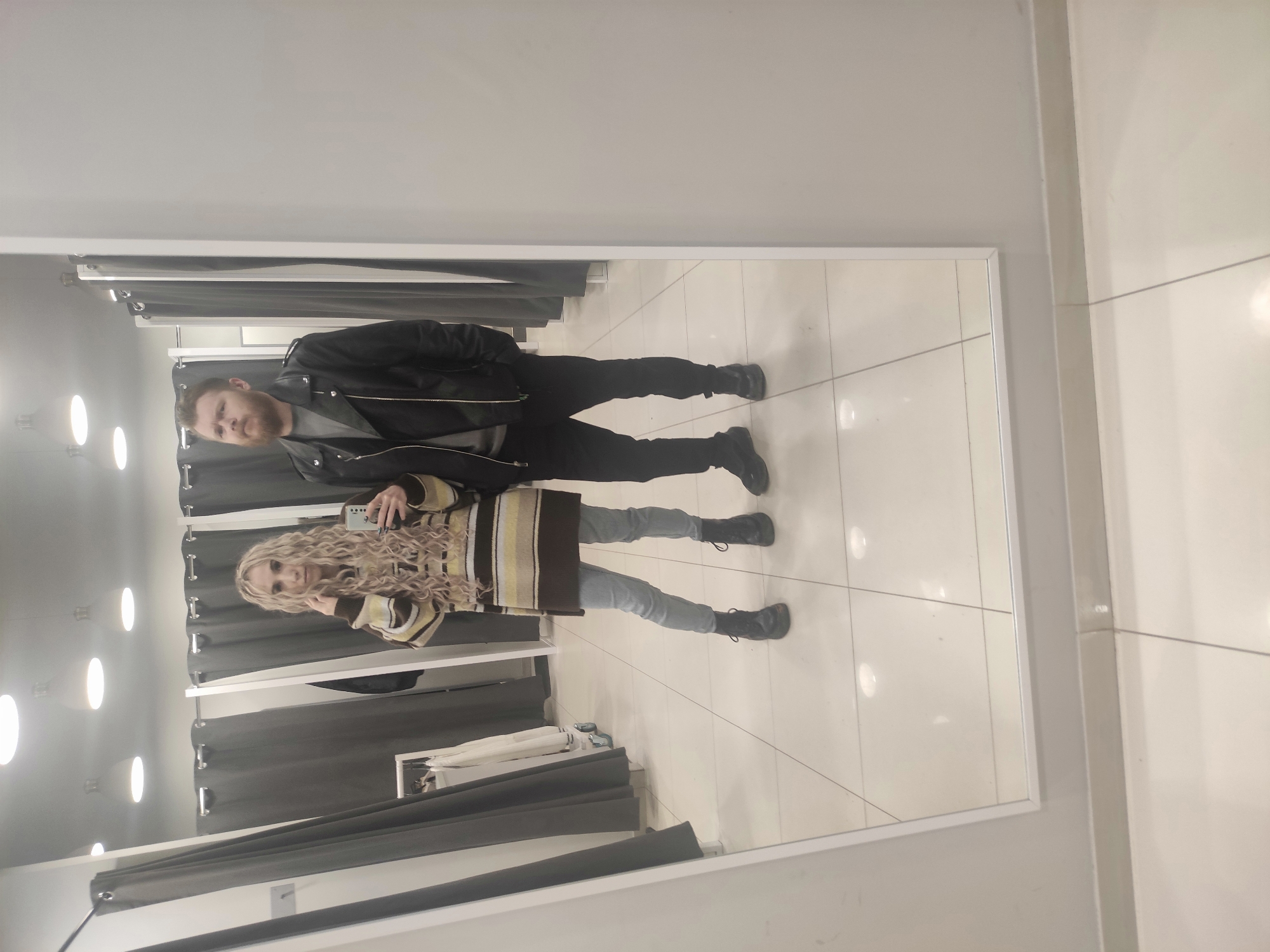Two individuals are reflected in a large mirror located within a fitting room in Chaikovsky, Russia. The room is characterized by a bright, white-tiled floor and white walls. Grey curtains hang along one side of the fitting room, obscuring racks with visible clothing. The male figure is clad in a black jacket, grey shirt, and black pants. The female figure wears a striped sweater, light grey pants, and is holding a mobile phone. The phone is presumably being used to capture the reflection of both individuals in the mirror.