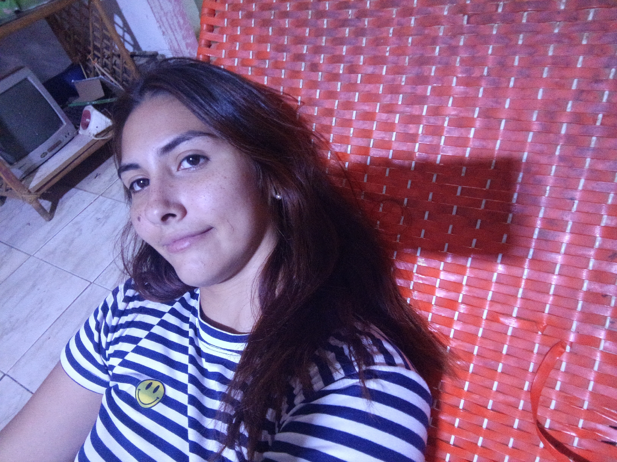 A young woman with dark hair and freckles is lying on a woven red mat, likely indoors, as evidenced by the interior background. She is wearing a striped blue and white shirt with a yellow smiley face patch. In the background, on the left, is an older television set on a wooden stand. Given the prompt, this scene is likely located in Juan Griego, Venezuela.