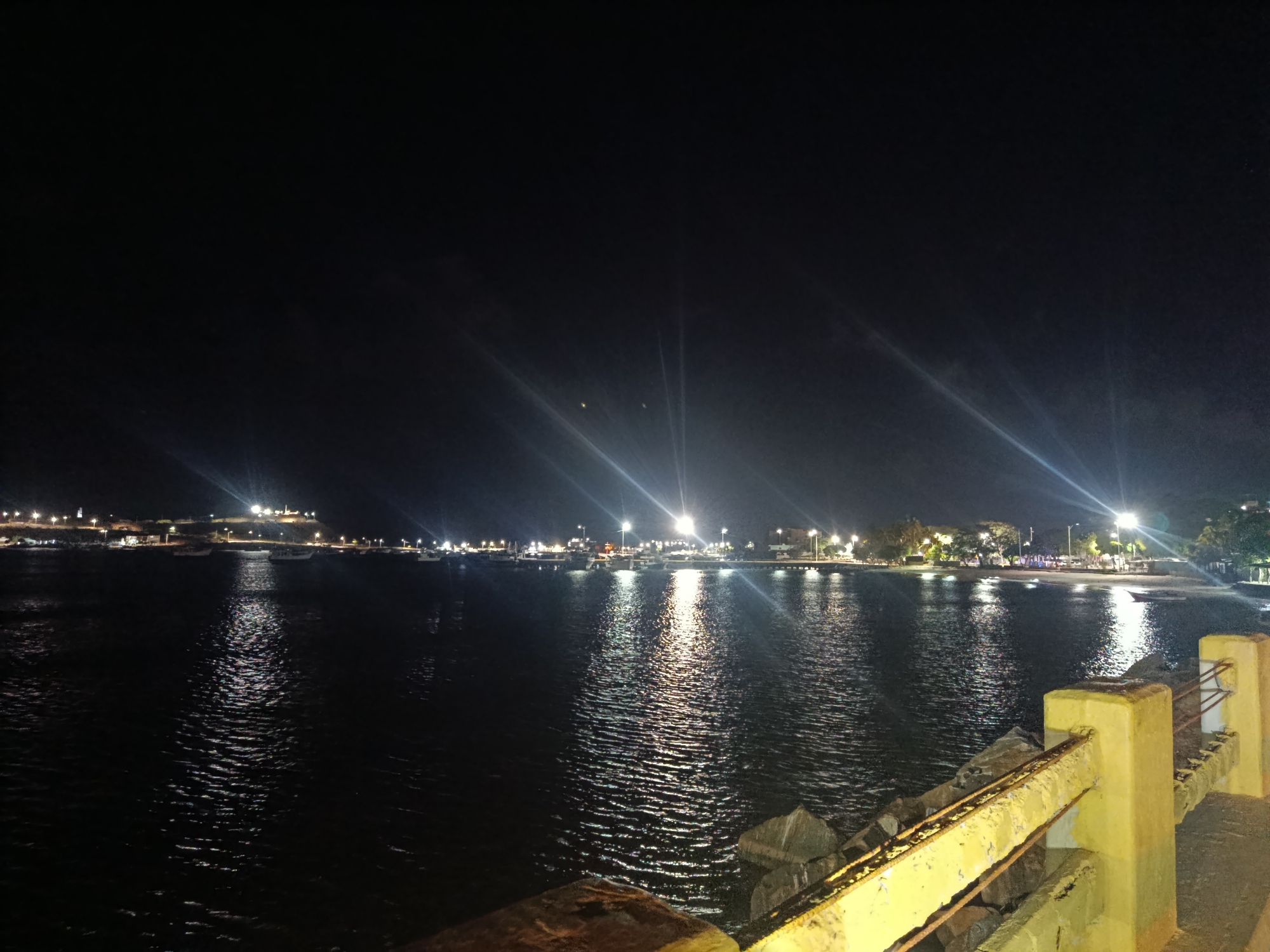 The image depicts a night view of the coastal city of Juan Griego, Venezuela. The foreground shows a yellow concrete barrier with corroded metal bars. Beyond the barrier, large rocks slope down to the water's edge. The dark water reflects the bright lights from the coastline. In the midground, the coastline is visible, illuminated by streetlights and lights from buildings. There are several boats docked in the harbor. In the background, a hill is seen, dotted with more lights. The sky is dark.