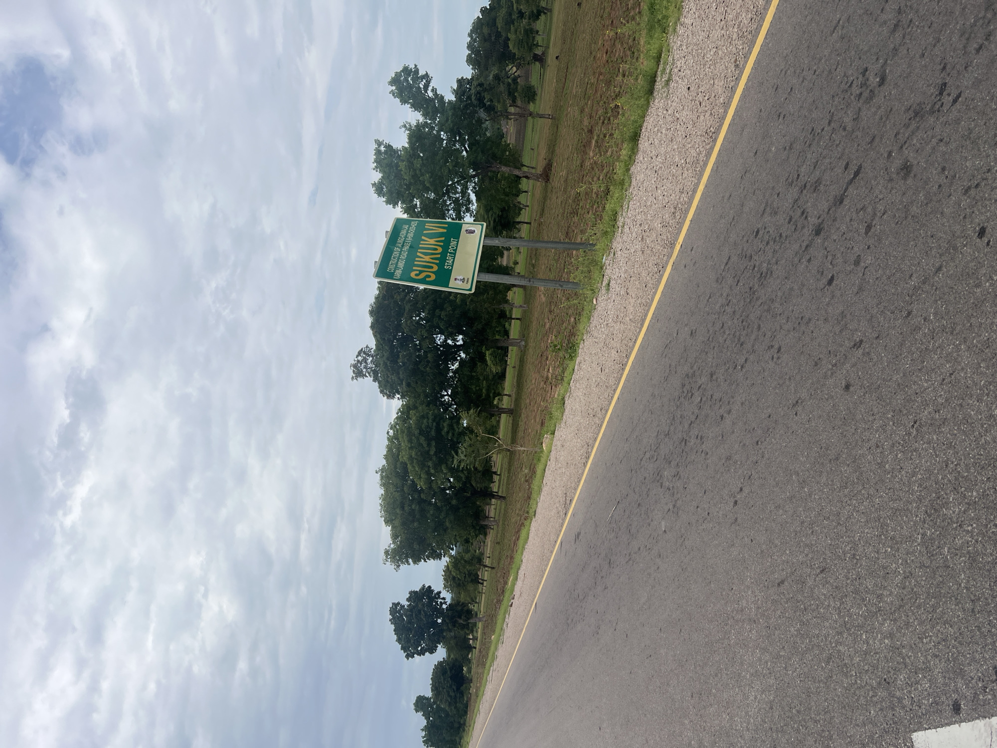 The image shows a roadside scene in Minda, Nigeria. The primary object of interest is a green signpost with white text indicating "SUKUK VI START POINT" and details about a construction project. The sign is placed on the side of an asphalt road with a yellow dividing line. Beyond the road, there is a grassy area with several large trees, some of which appear to be mature. The background consists of a cloudy sky. The overall scene suggests an outdoor, daytime setting, likely in a rural or semi-rural area. There are no people visible in the image. The weather appears overcast, with a cloudy sky dominating the upper portion of the frame. The text on the sign provides specific information about a road construction project, indicating the beginning of phase six of the Jalino-Kona-Lau Karim-Lamido Road project.