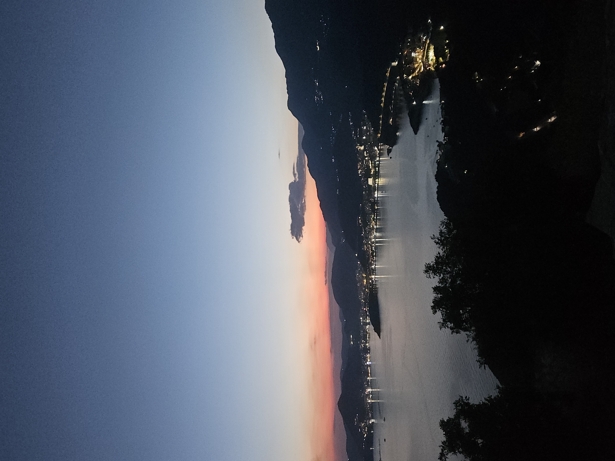 The image shows a twilight view of Sveti Stefan, Montenegro, from an elevated viewpoint looking towards the coast. The sky transitions from a deep blue at the top to a soft orange and pink gradient near the horizon, indicating the sun has recently set. Silhouetted against the colorful sky are the dark outlines of hills and mountains.
Below the sky, the town of Sveti Stefan is visible, with numerous lights twinkling along the coastline and up the hillsides. The lights suggest a populated area with buildings and infrastructure. A body of water, likely the Adriatic Sea, occupies the middle ground, reflecting the ambient light. The water appears calm with a few subtle ripples.
In the foreground, the dark silhouettes of trees and foliage obscure the ground, suggesting the viewpoint is from a natural area overlooking the town. The overall scene captures the serene beauty of a coastal town at dusk. There are no discernible people or specific actions depicted in the image. The environment is characterized by natural elements like the sea, mountains, and vegetation, as well as the man-made lights of the town.