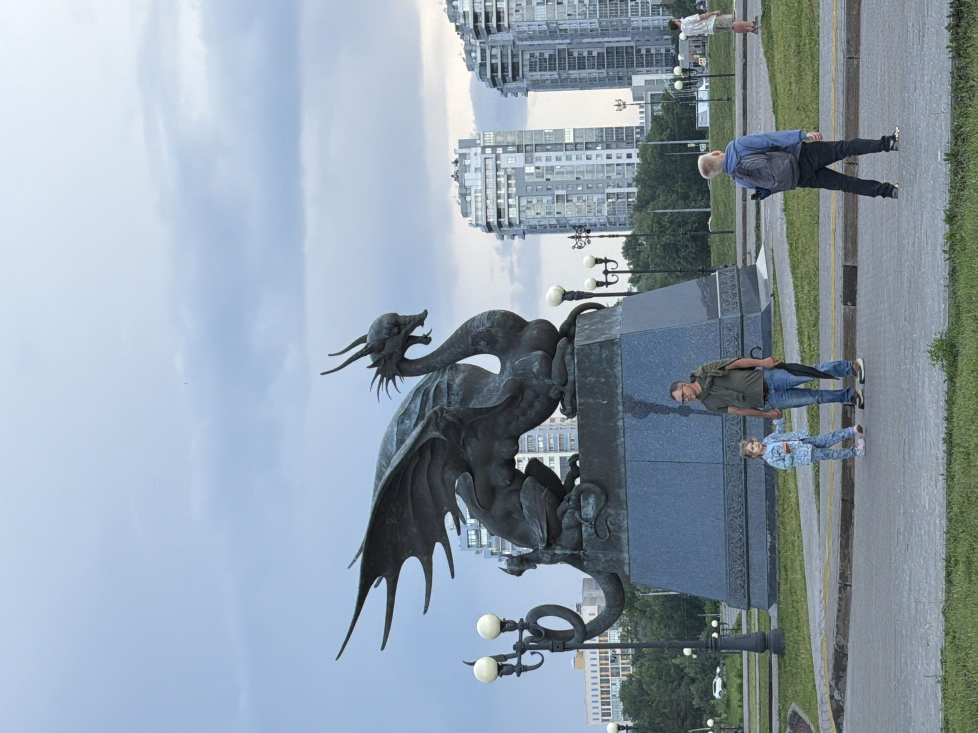 A bronze sculpture of a dragon is prominently featured in a city park, likely in Kazan, Russia, given the presence of the stylized architecture in the background. The dragon, with its intricate wings spread and head turned to the left, rests on a large, dark granite pedestal. In the foreground, a man in a green t-shirt and jeans stands next to a young girl in a denim jacket and pants. The girl is holding a small toy. To the right of the man and girl, a young boy with a blue backpack walks away from the camera. In the background, modern high-rise buildings with reflective windows rise against a cloudy sky. The scene is set in an urban park with manicured grass, paved walkways, and decorative lampposts.