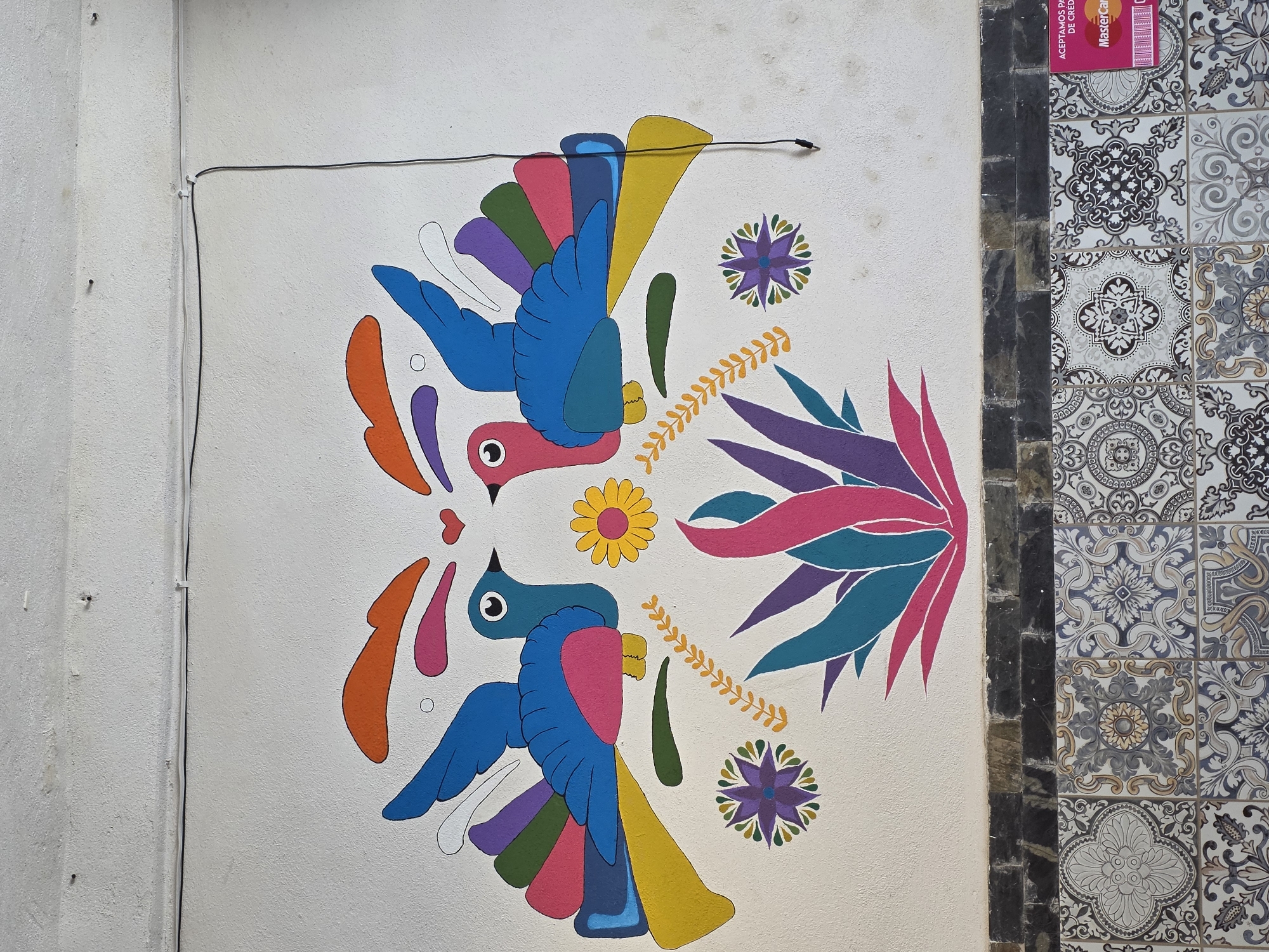 A close-up shows a wall in Puebla, Mexico, adorned with a vibrant mural. The mural features two mirrored, stylized blue birds with pink heads, accompanied by colorful elements such as flowers, leaves, and ornamental shapes in shades of orange, purple, green, and yellow. A black electrical wire runs across the top of the mural. To the right of the painted wall is a section covered in decorative ceramic tiles with intricate grey and white patterns, separated by a column of dark stone. A red credit card advertisement is visible on the top of the tile section.