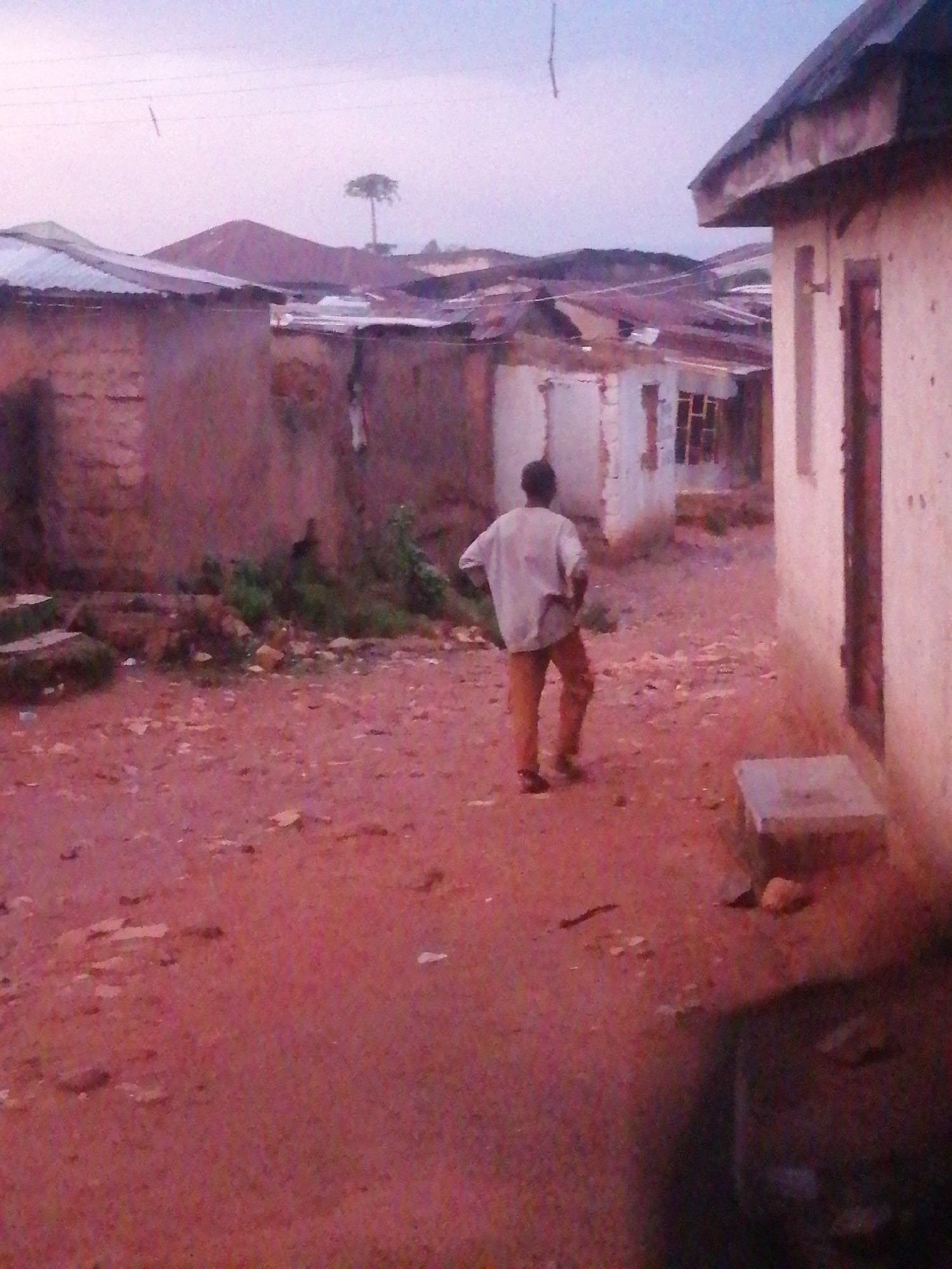 A solitary male, seen from behind, walks down a dirt path in an impoverished urban area. He wears a light-colored, long-sleeved shirt and tan pants. The path is a reddish-brown, dusty surface scattered with small stones and debris. To his left, buildings constructed with rough, light-colored brick walls and corrugated metal roofs are visible. Some of these structures appear dilapidated, with exposed brickwork and uneven roofing. To his right, a building with a cream-colored exterior and a dark, possibly wooden, doorway stands. A concrete structure, possibly a bench or a platform, rests on the ground to the right of the path. In the background, more buildings with similar roofing materials are clustered together, and a solitary, tall tree with a sparse canopy stands against a pale, overcast sky. The overall impression is one of a rural or informal settlement within a city.