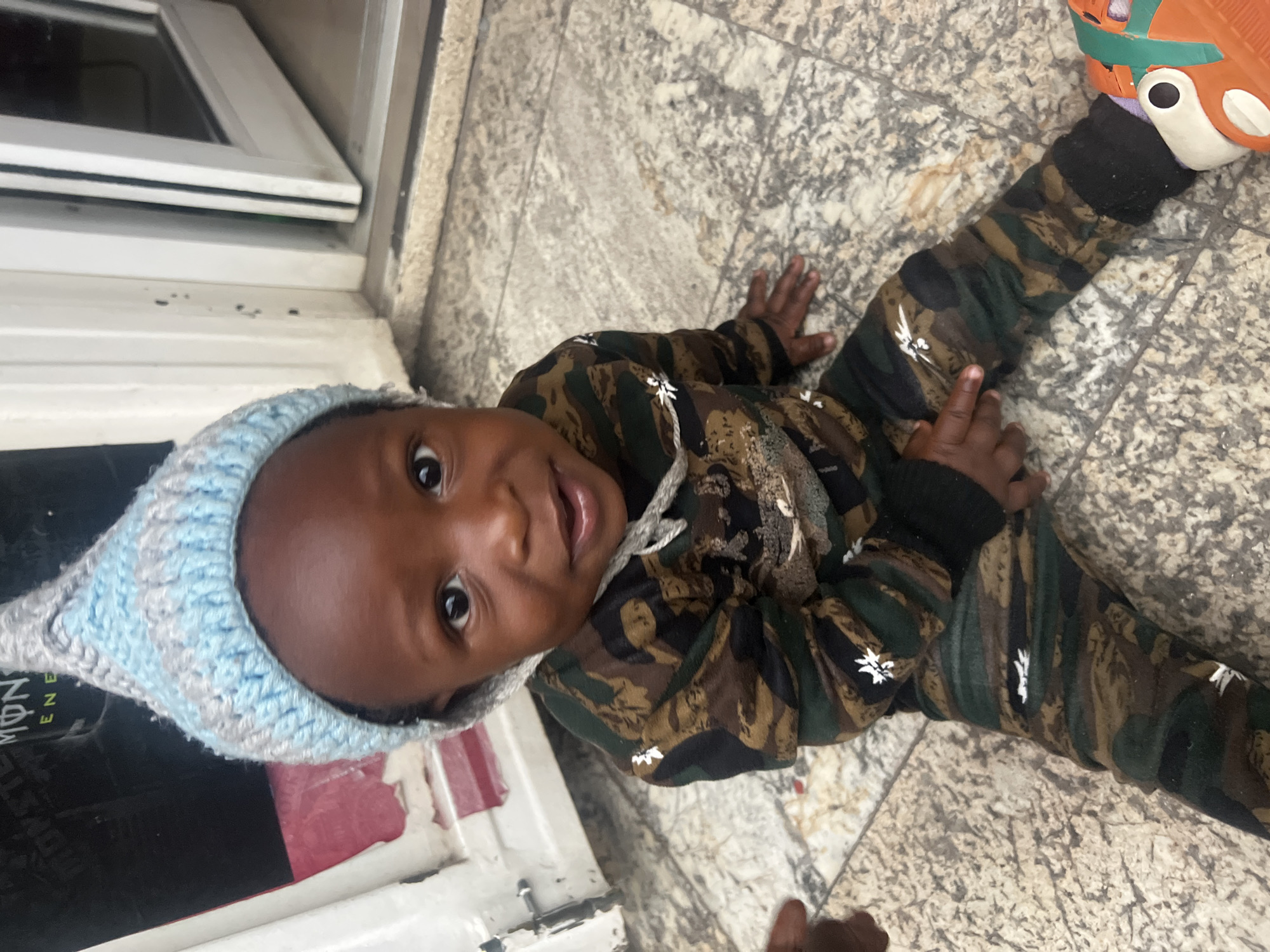 A young child wearing a blue and grey knitted hat and camouflage patterned clothing lies on a tiled floor next to a white painted window frame. The child is looking directly at the camera. One of the child's feet is covered by a bright orange shoe. It is not possible to confirm the location as Gura Top, Nigeria based on the given image.