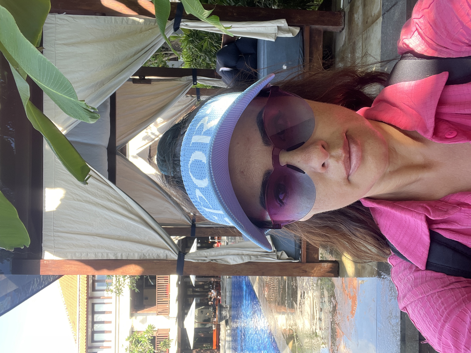 A woman, possibly of South Asian descent, is taking a selfie outdoors, wearing a pink, collared shirt and a light blue visor with white lettering. She has on large, purple-tinted sunglasses. Her long, dark brown hair is visible. The background suggests a resort or hotel setting, with a swimming pool, lounge chairs, and thatched-roof cabanas. The scene is brightly lit, indicating it is daytime, and the weather appears to be warm and sunny. The woman's expression is relaxed, with her mouth slightly open. The environment suggests a tropical location, consistent with the provided location context of Kuta Selatan, Indonesia.