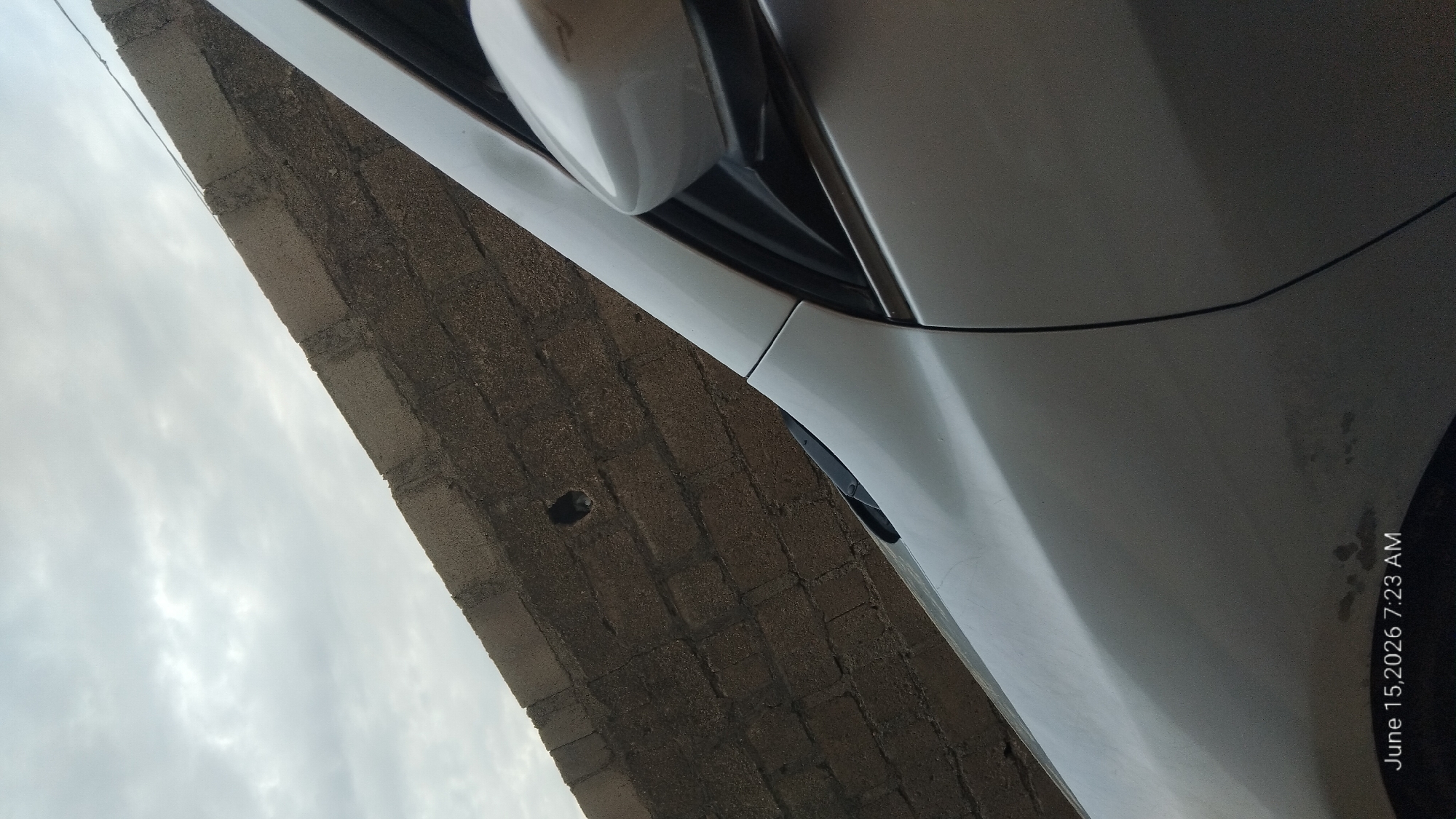 The image shows a close-up view of a silver car parked next to a rough, textured brick wall. The wall appears weathered and has a dark, mottled appearance. The sky above is overcast with light gray clouds. The car's side panel and side mirror are visible, reflecting the muted light. A small, dark circular imperfection is noticeable on the brick wall. The timestamp in the lower left corner reads "June 15, 2026 7:23 AM." There are no people or discernible actions taking place in the scene. The overall impression is that of an outdoor urban or semi-urban environment.
