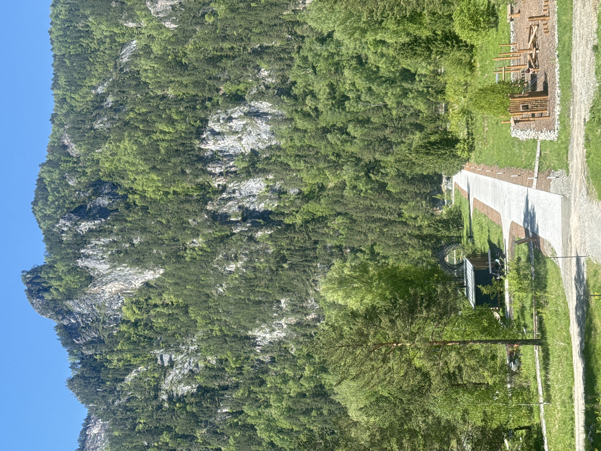 The image depicts a landscape scene with a mountain dominating the left side of the frame, covered with dense green trees and rocky outcrops. A clear blue sky is visible above the mountain. To the right, a path or road leads towards a small building or structure with a metallic roof. Nearby, there is a grassy area with a wooden playground structure. A large tree is positioned prominently in the foreground, partially obscuring the view of the building.
The image does not contain any people, actions, or interactions, only objects and the environment. The provided location of Ust'-Muny, Russia, cannot be confirmed without additional information or context within the image.