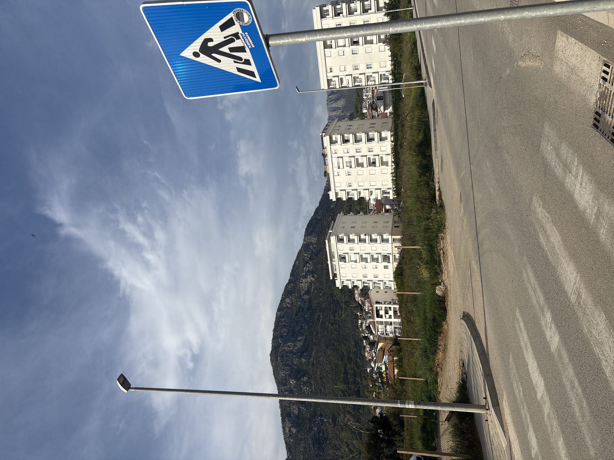 The image shows a street in Bar, Montenegro. In the foreground, there is a blue and white pedestrian crossing sign on a metal pole. Behind the sign, a road with white stripes runs alongside a grassy area. Several tall, white apartment buildings are visible in the background, situated at the base of a large, rocky mountain. A street lamp is seen on the left side of the frame, reaching towards the sky which is lightly covered with clouds.