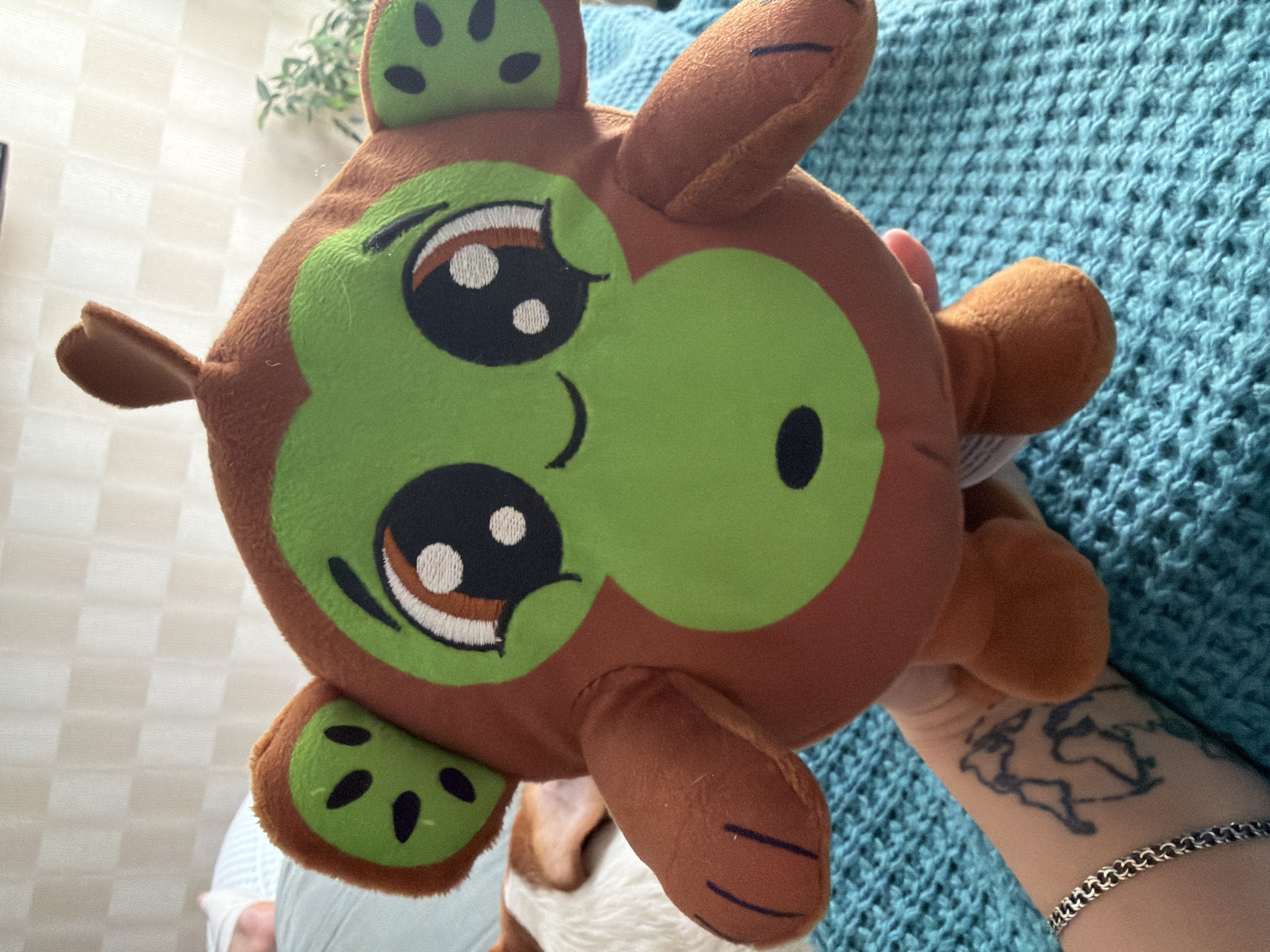 A close-up shot focuses on a round, plush monkey toy with a green face and brown fuzzy ears patterned like kiwi slices. The toy has large, expressive embroidered eyes with brown and white irises and black pupils. Its mouth is a simple curved line, and a small oval is embroidered on its green cheek. The monkey is held by a hand that is visible at the bottom left of the frame, adorned with a silver bracelet and a dark tattoo on the wrist. In the background, a textured teal-colored blanket is visible, along with the hindquarters of a dog with white and tan fur. A glimpse of a white pillow and a person's leg is visible in the upper left background, along with a blurred green plant and a wall with a checkered pattern. The scene appears to be indoors.
