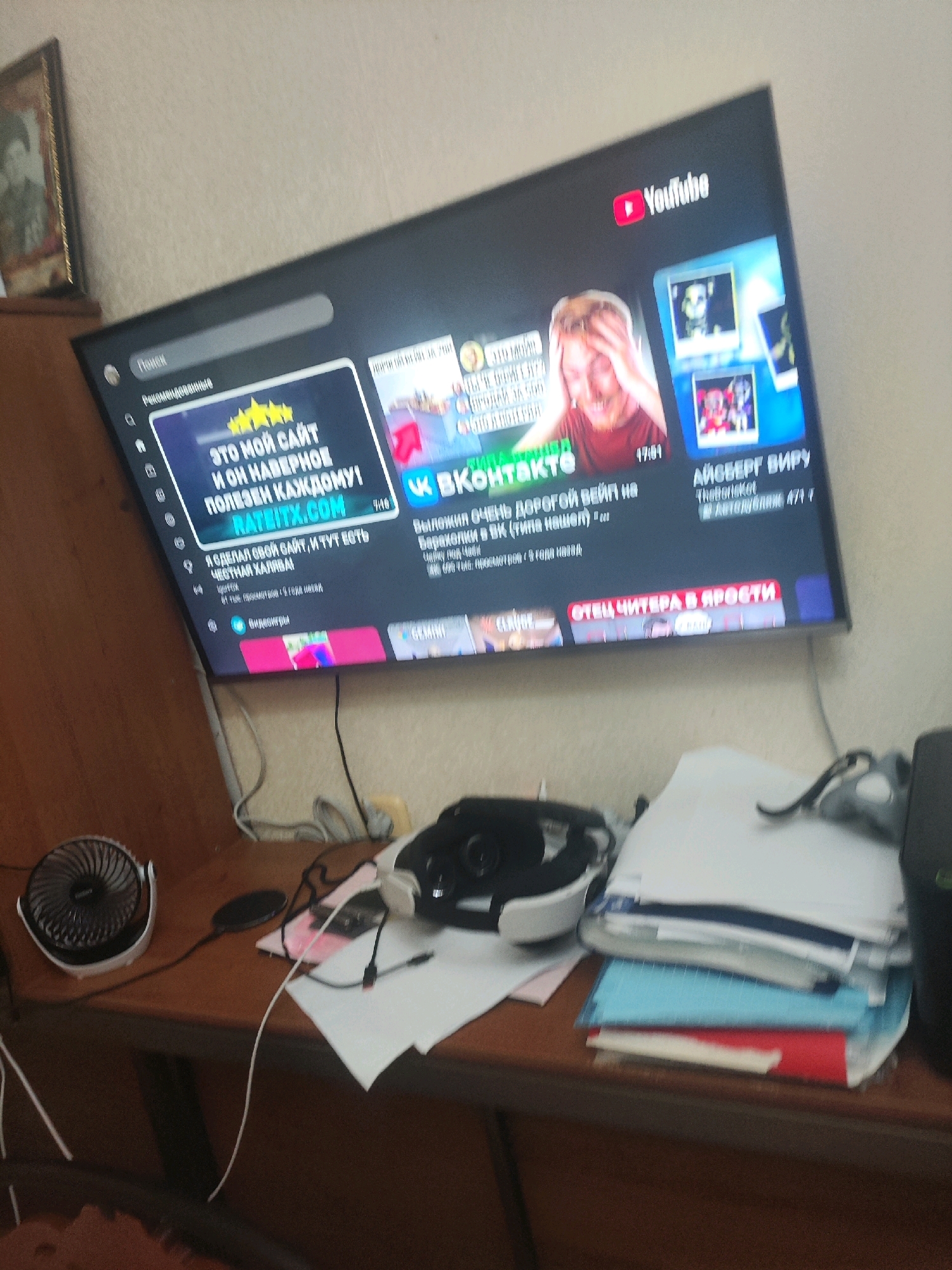 The image shows a large television screen displaying the YouTube interface. The screen is mounted on a wall and is the central focus of the image. On the left side of the screen, a search bar and a "Recommended" section are visible. A prominent advertisement on the screen features text in Russian, suggesting it is a Russian-language platform. The advertisement includes images of people and promotional graphics. The television is positioned above a wooden desk cluttered with various items. These items include a small black fan, a charging pad, a virtual reality headset, stacks of paper, and other electronic devices. In the background, a framed photograph is visible on the wall to the left of the television, and the overall environment appears to be a domestic or office setting. The lighting in the room is moderate, with some ambient light contributing to the scene's illumination. The image is a still photograph taken from a slightly elevated angle, capturing the television screen and the immediate surroundings.