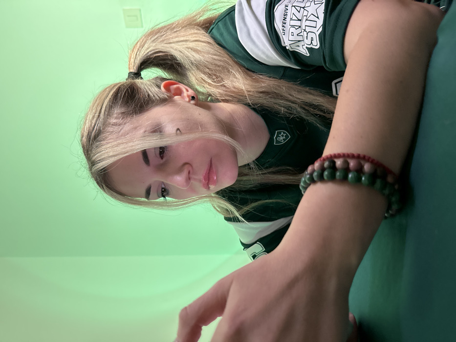 In Budva, Montenegro, a blonde female is depicted in an indoor setting. She wears a dark green long-sleeved shirt with white stripes, displaying "OFFENSIVE ARIZONA STATE" text on the right sleeve and a shield-like logo on the upper left chest. Her hair is pulled back into pigtails, with some strands framing her face. A small, dark crescent-shaped marking is visible on her right cheek below the eye, and she has a dark stud earring in her right ear. Her eyes are directed downwards and slightly to her left, consistent with looking at an object held by her partially visible right hand, which is out of focus in the upper left. Her left arm rests on a dark green surface, wearing a beaded bracelet composed of green and reddish-brown stones, layered over a red braided string bracelet. The background is uniformly illuminated with a light green color, featuring a room corner and a white wall fixture on the right.